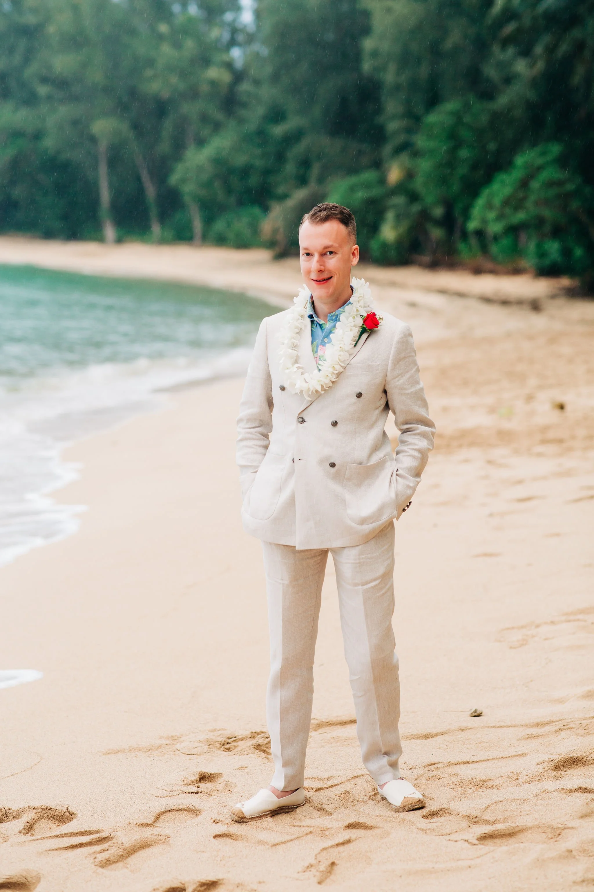 A man in a cream-colored, double-breasted suit with a floral shirt stands on a sandy beach with greenery in the background. He is wearing a lei and a red flower on his lapel, and has his hands in his pockets, smiling at the camera.