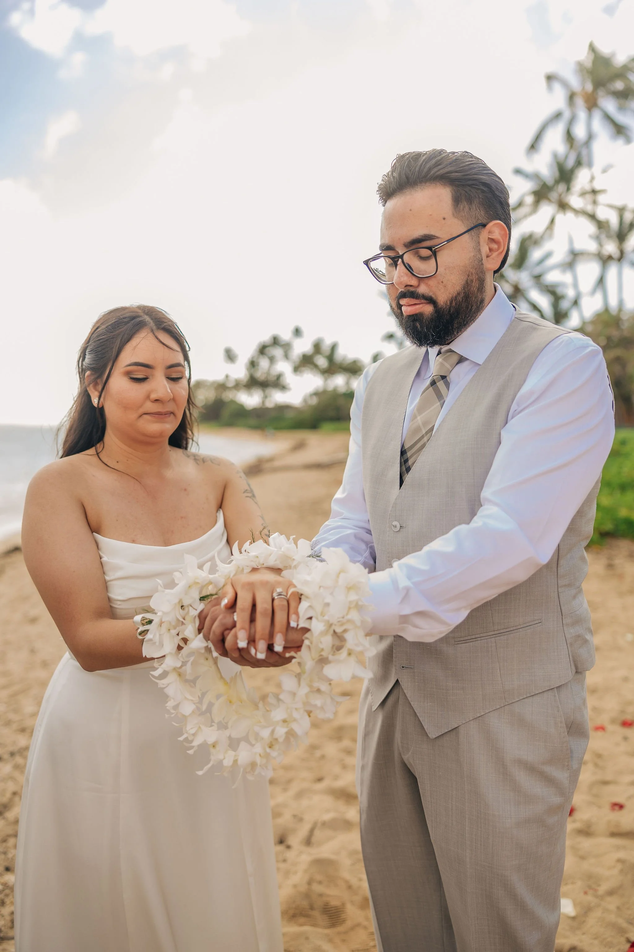 A couple dressed in wedding attire exchanging rings on a beach, with a cloudy sky and trees in the background.