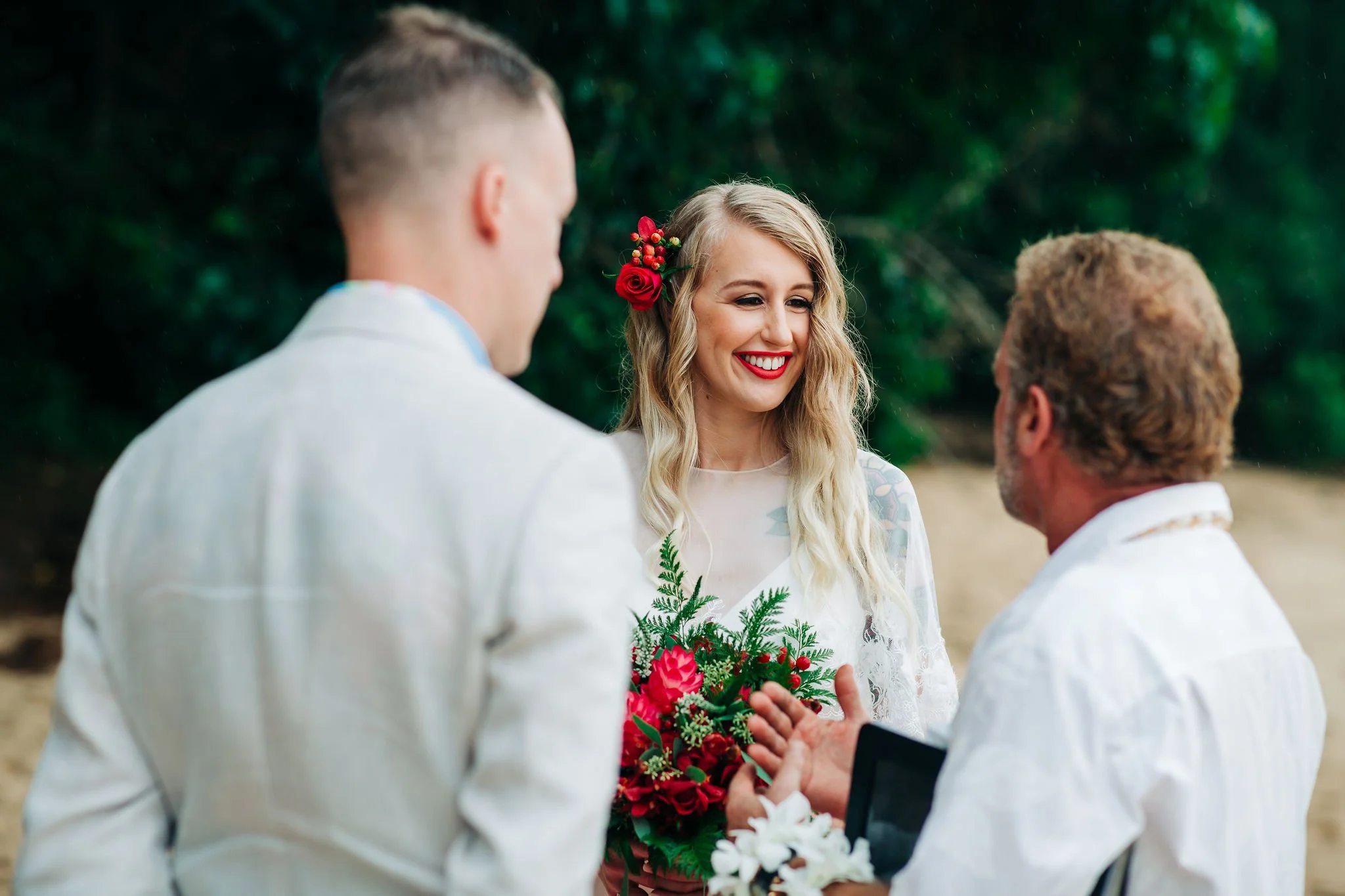 A wedding ceremony outdoors with a woman holding a bouquet, smiling, with two men in white shirts, one with his back to the camera, the other facing her and gesturing.