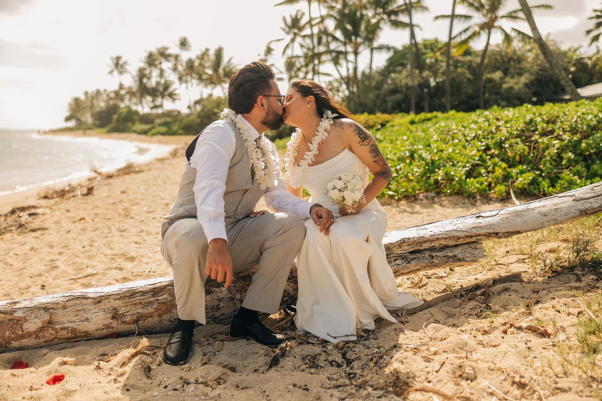 A bride and groom sitting on a log on a sandy beach, kissing, with tropical trees and the ocean in the background, during their wedding.