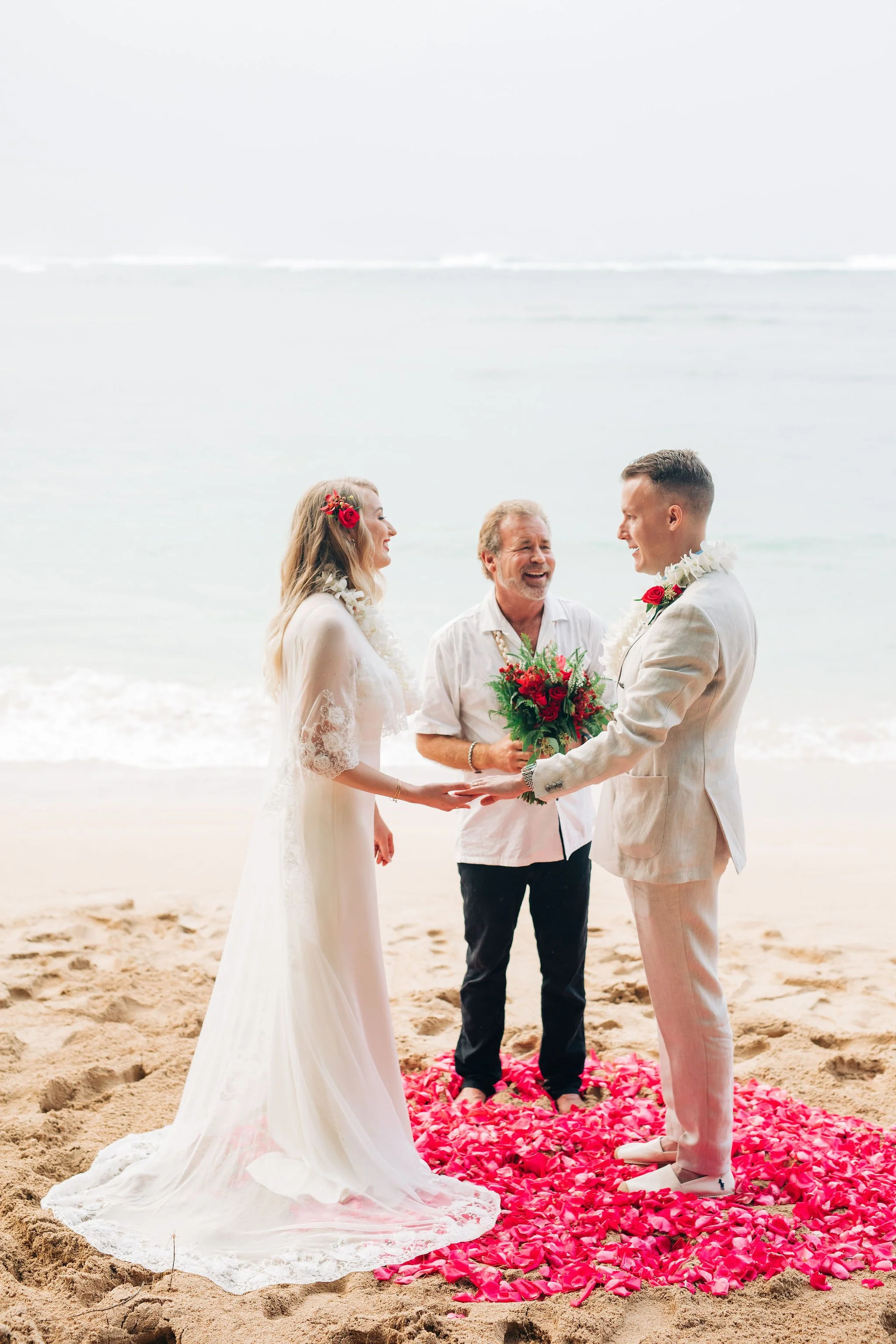 A couple getting married on the beach with an officiant, standing on a bed of pink flower petals, with the ocean in the background.