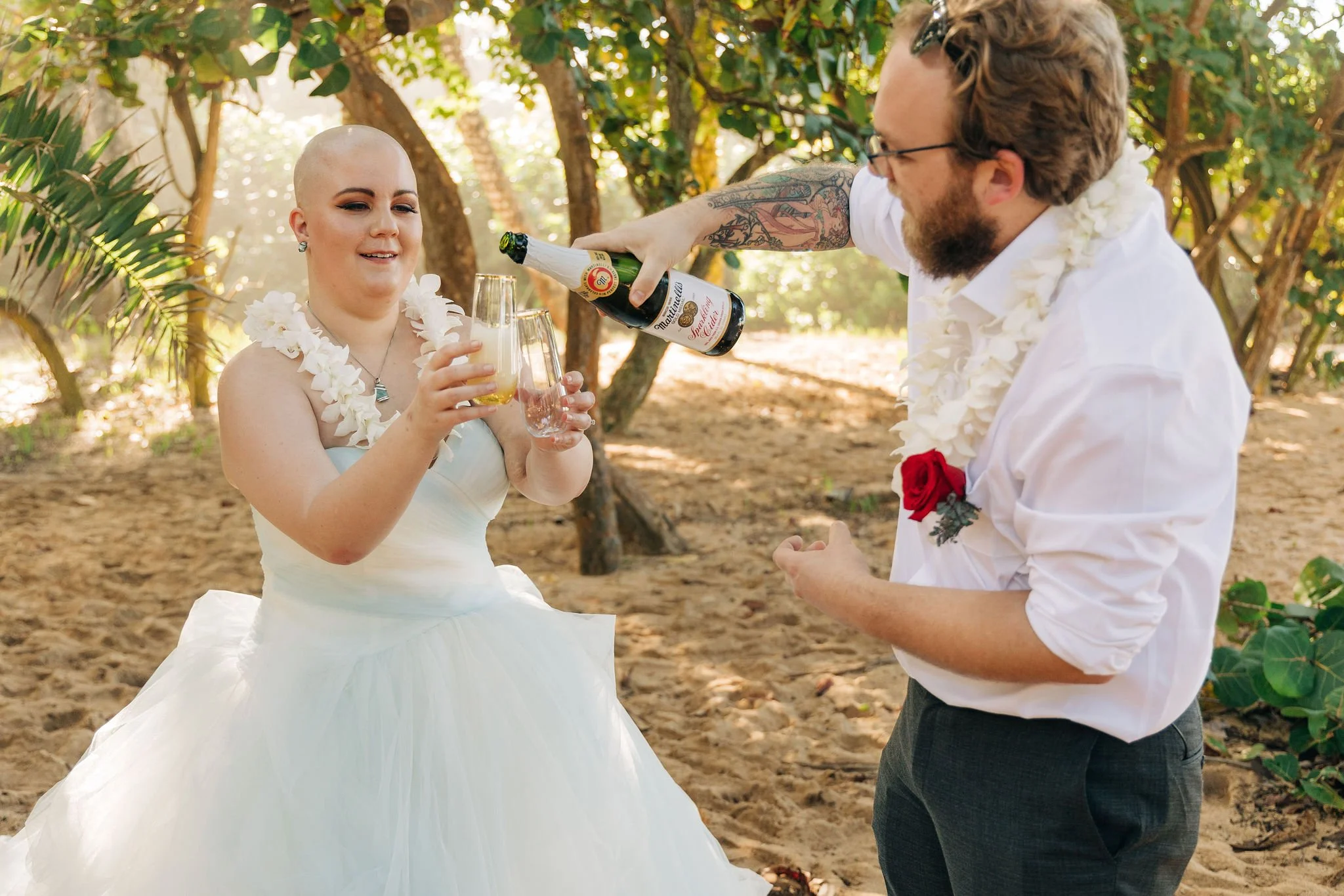 A couple dressed in wedding attire toasting with drinks during an outdoor wedding ceremony on a sandy beach with green trees in the background.