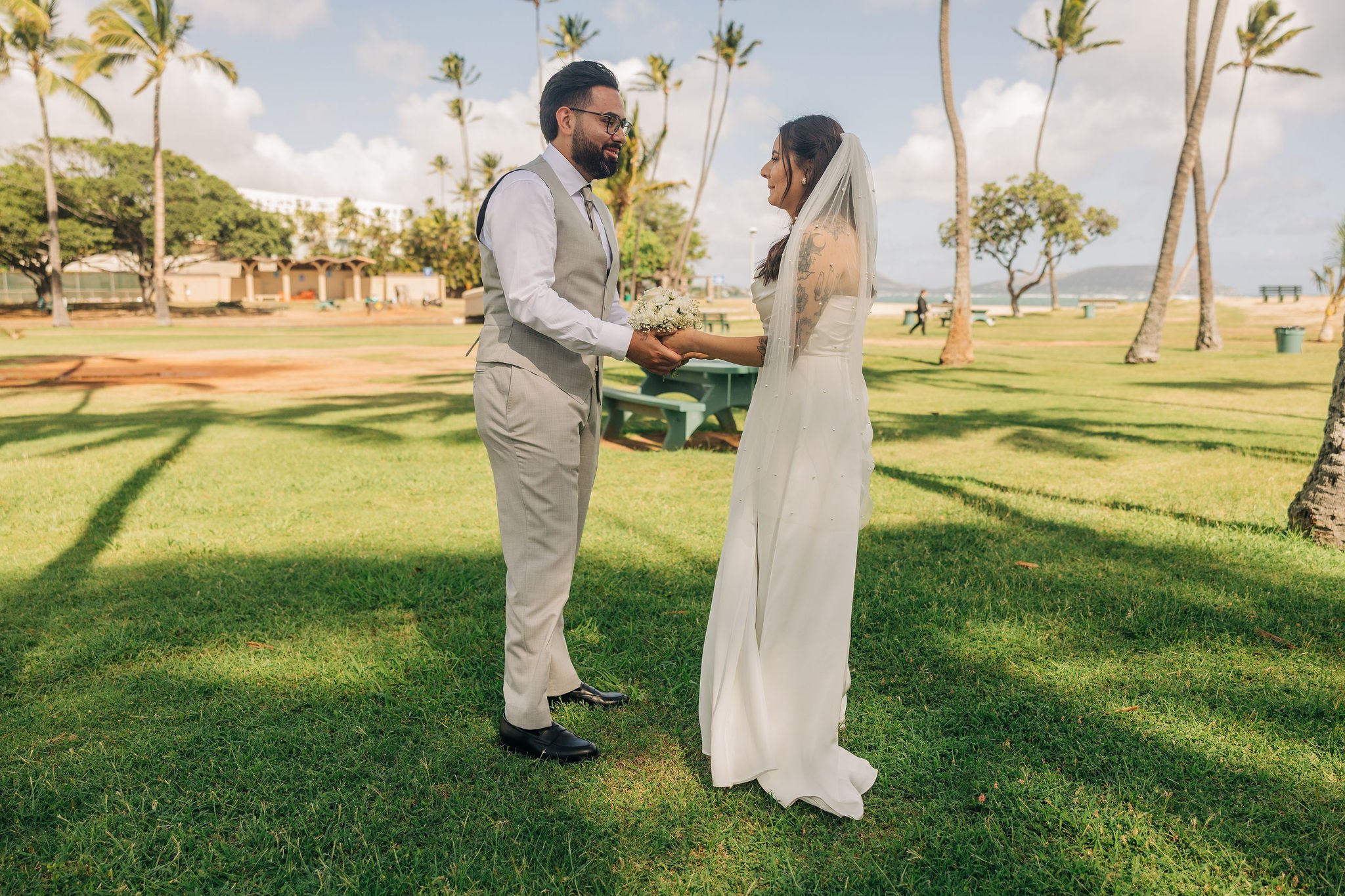 Couple exchanging wedding vows outdoors on a grassy area with palm trees, a beach, and a pavilion in the background.