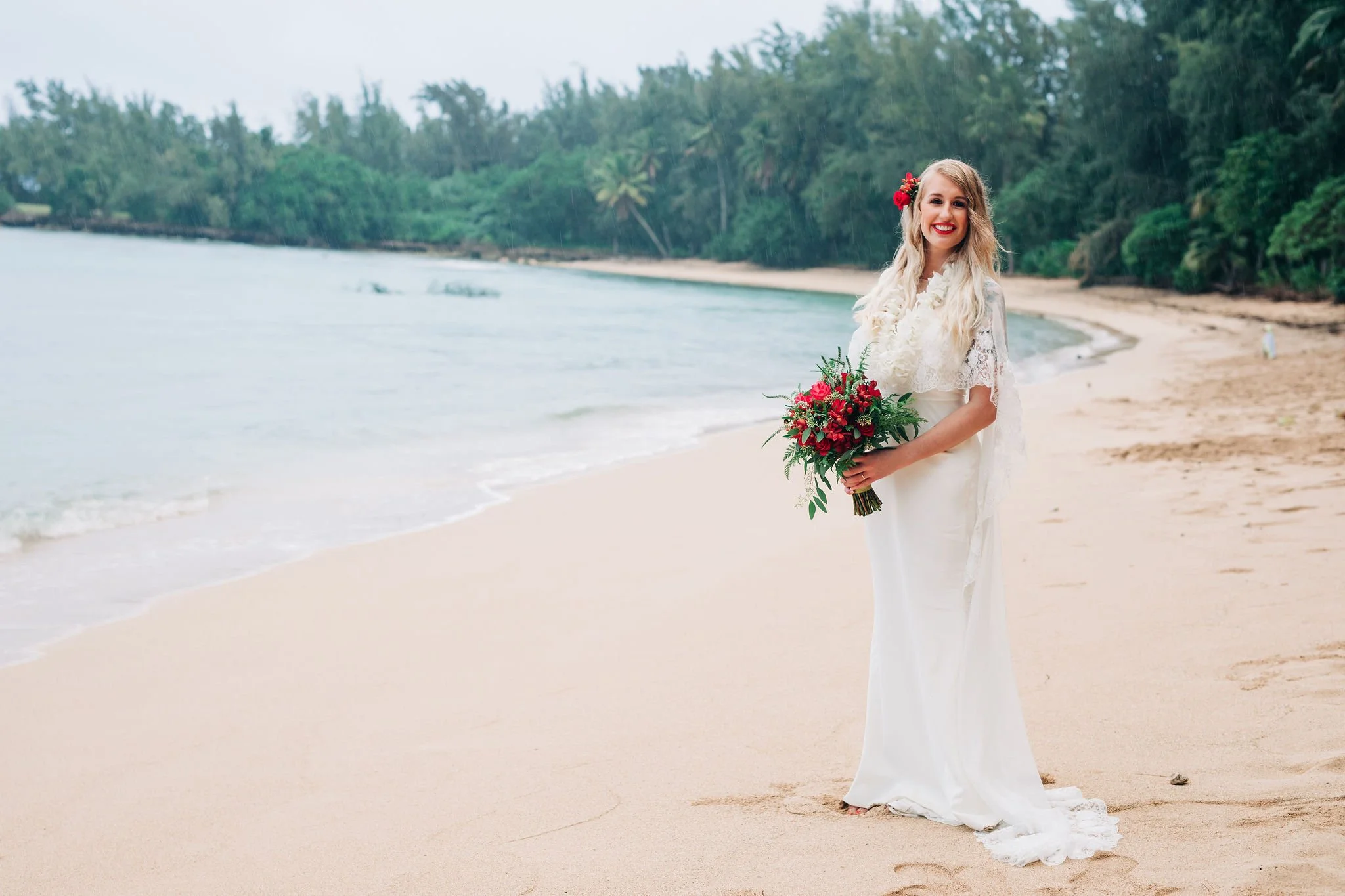 A bride in a white wedding dress holding a bouquet of red and green flowers, standing on a sandy beach with the ocean and a lush green forest in the background.