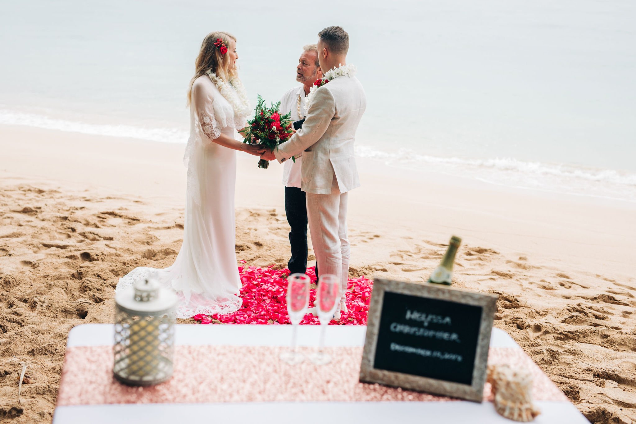 A wedding ceremony taking place on a beach with the couple exchanging vows. The bride and groom are holding hands and standing on a pink flower petal aisle, with a officiant in between them. The bride is wearing a white wedding dress, and the groom i