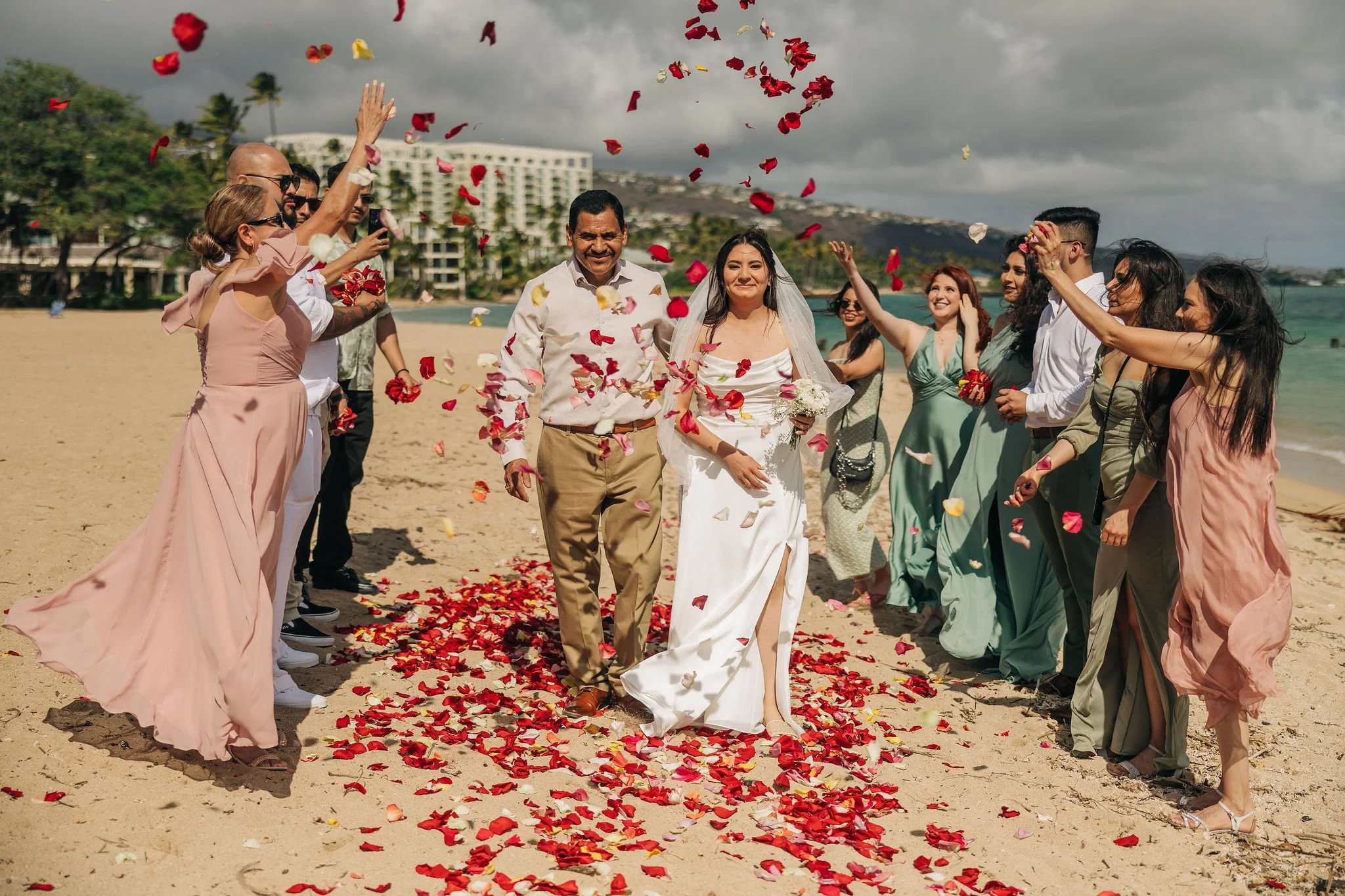 A bride and a man walking on a wedding ceremony setting on the beach, surrounded by friends throwing flower petals in the air, with a cloudy sky and buildings in the background.