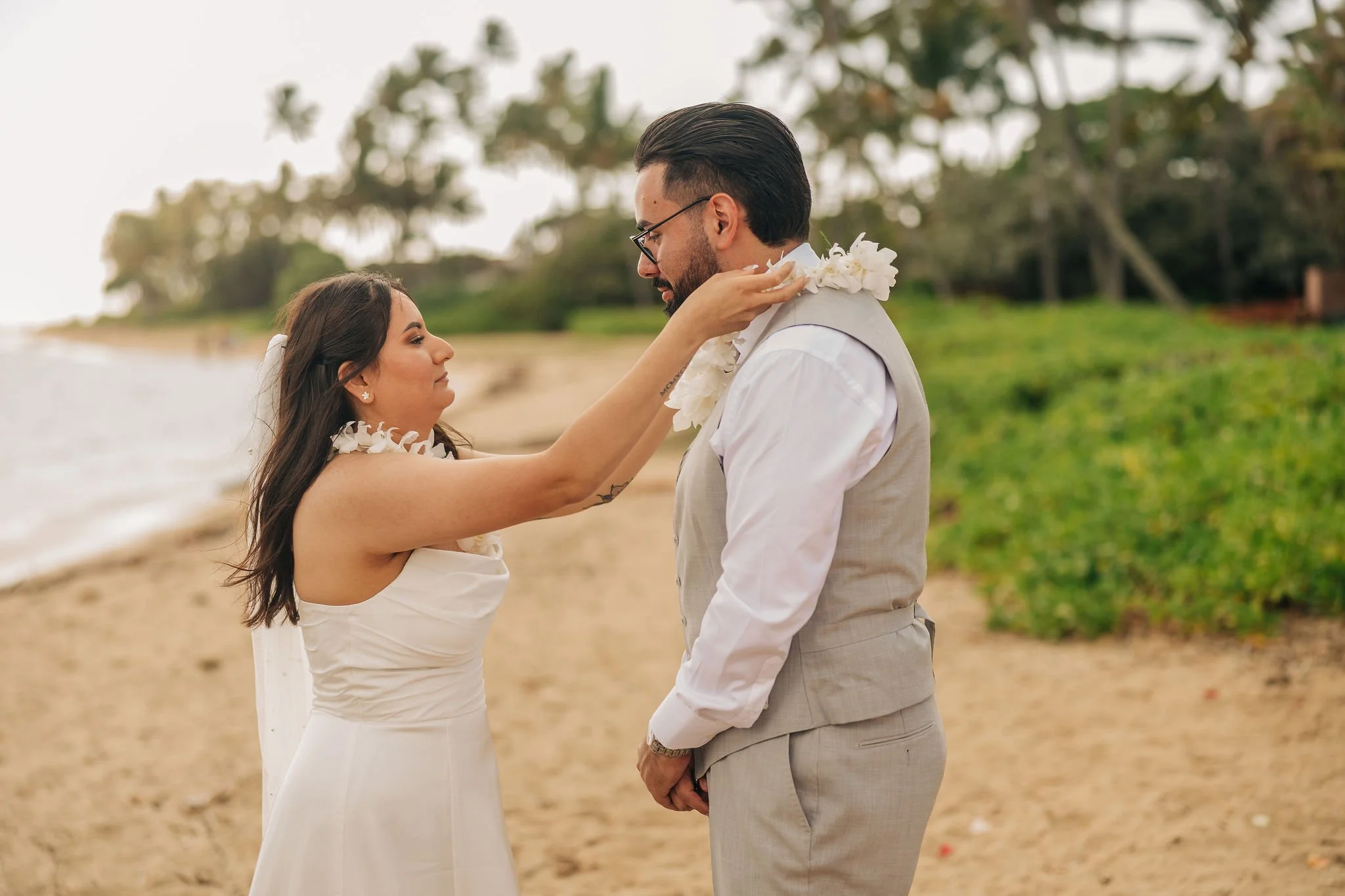 A woman in a white dress places a lei around a man's neck on a beach, with trees in the background.