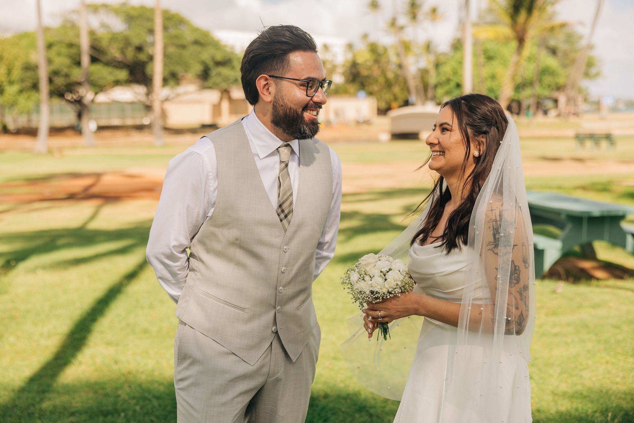 A bride and groom smiling at each other outdoors, with the bride holding a bouquet of white flowers and wearing a veil, and the groom in a beige vest and tie, in a park setting.