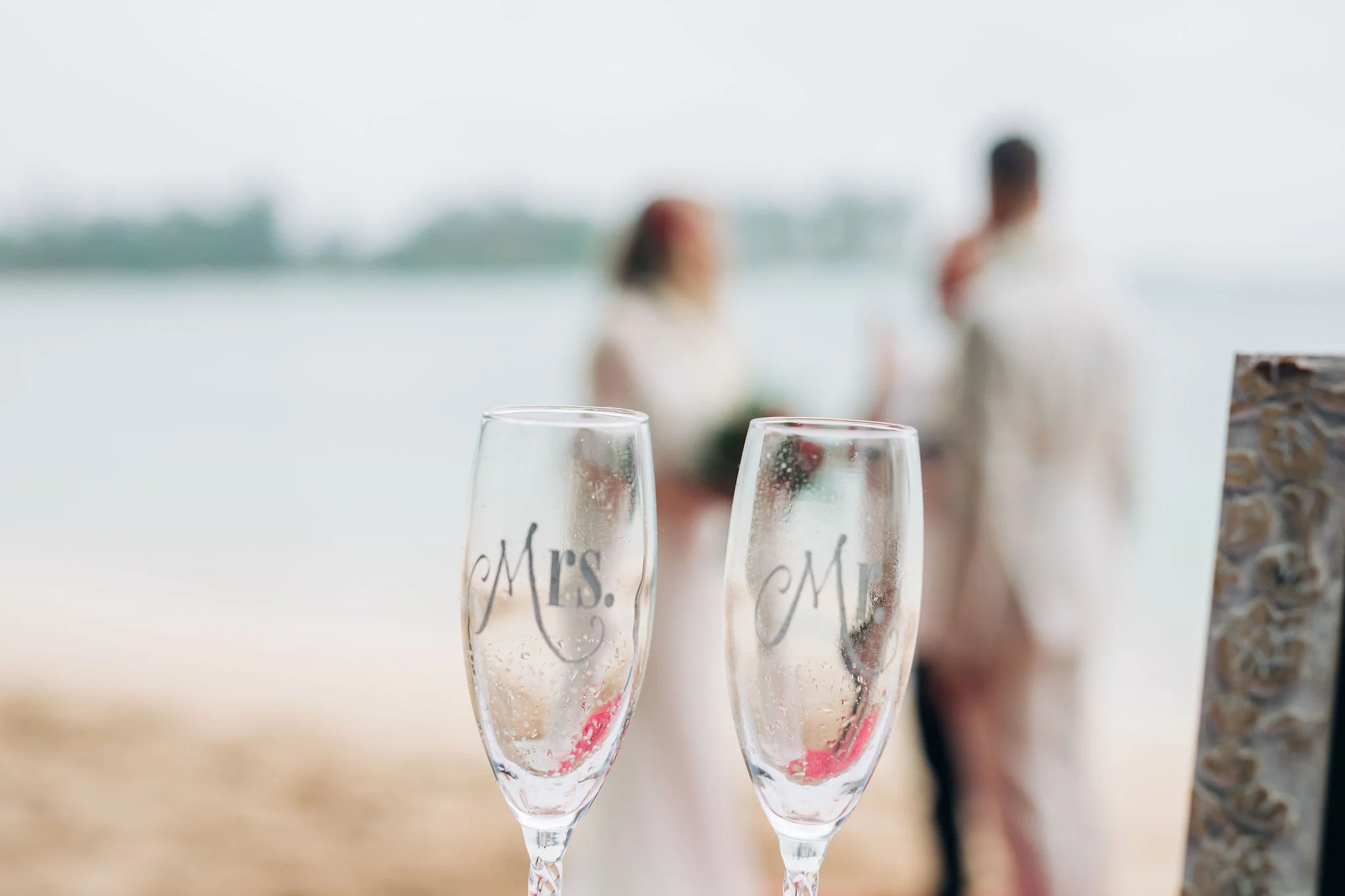 Two champagne glasses with 'Mrs.' and 'Mr.' written on them, blurred couple in wedding attire in the background at the beach.