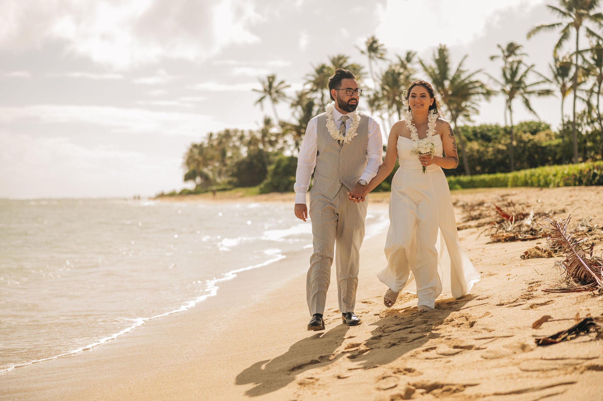 A bride and groom walking hand in hand on a beach, dressed in wedding attire, with palm trees and a cloudy sky in the background.