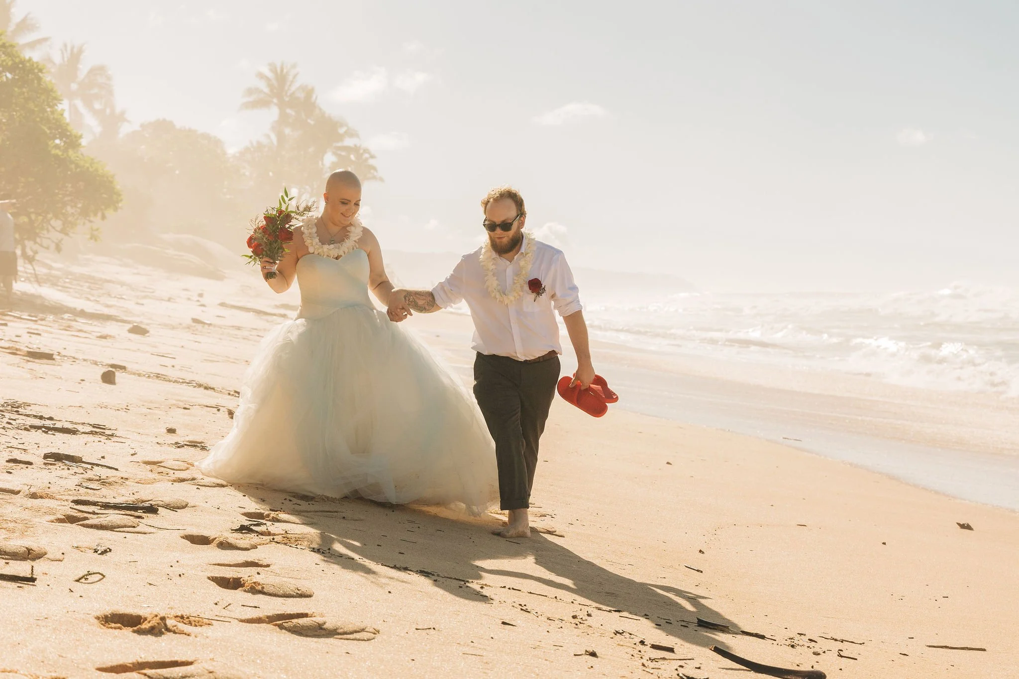 A bride and groom walking barefoot on the beach while holding hands, with the bride holding a bouquet and the groom carrying red shoes, under a bright sky with palm trees in the background.