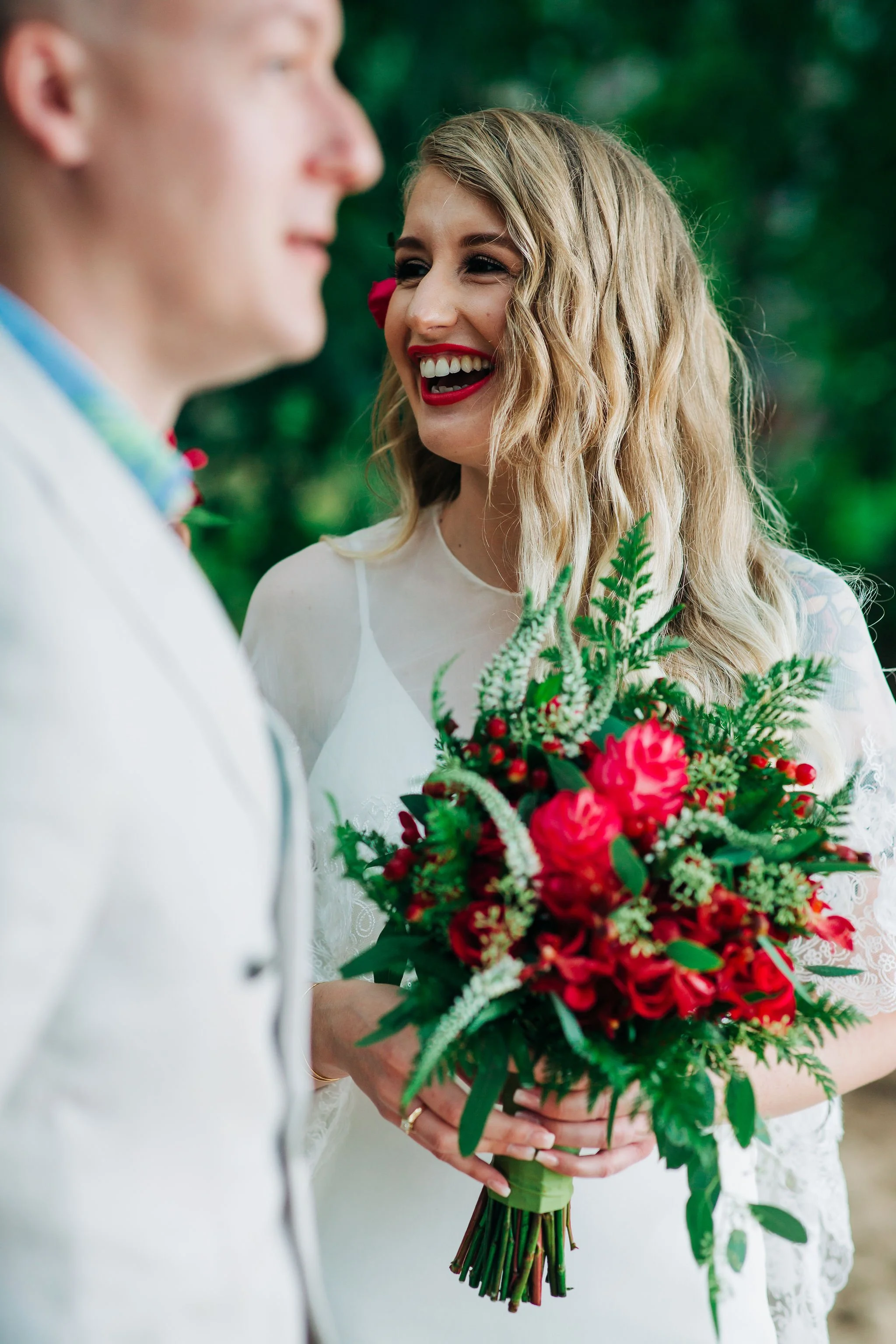 A woman in a white dress holding a vibrant red and green bouquet, laughing joyfully at a man dressed in white during an outdoor wedding.