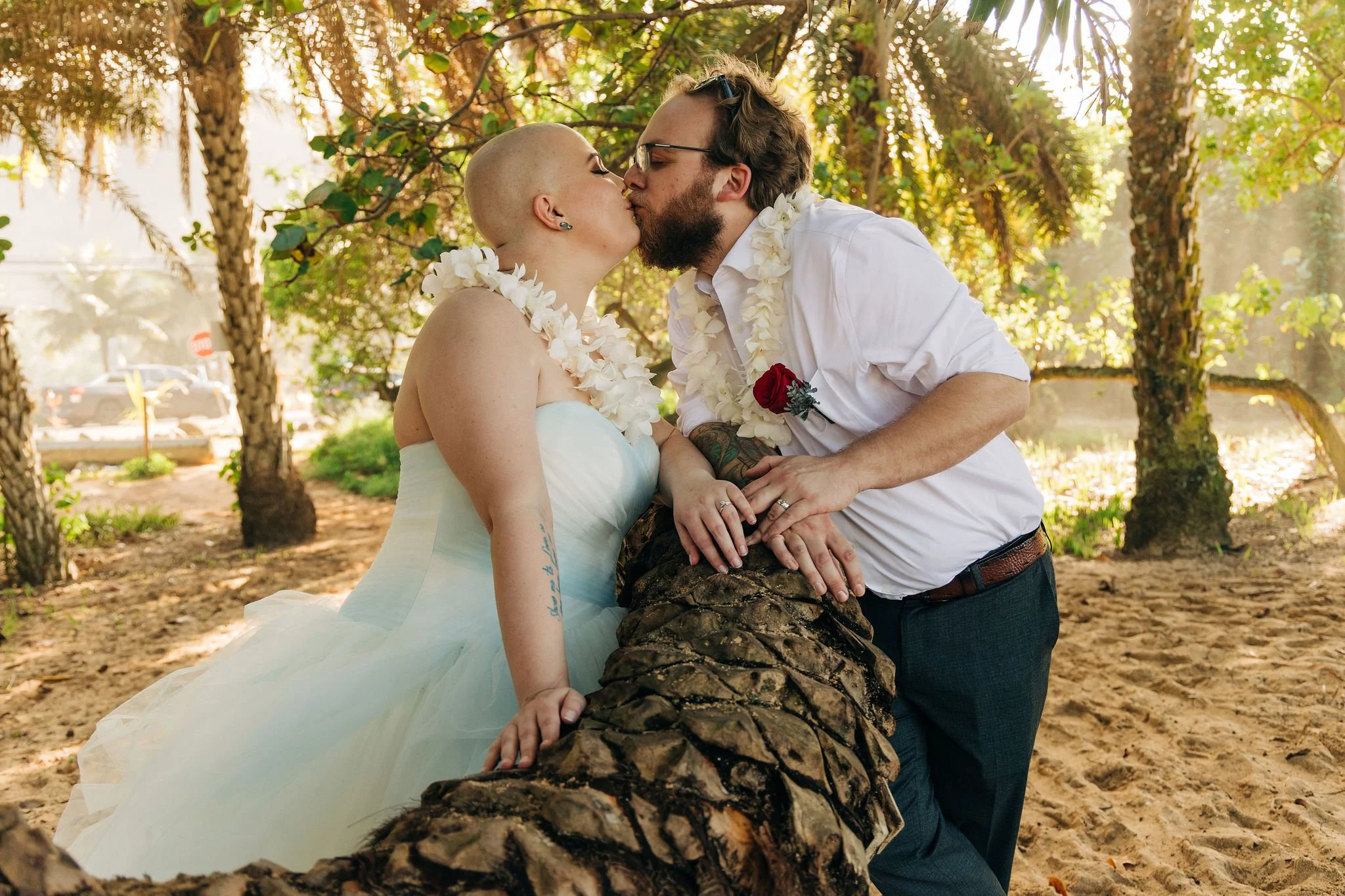 A same-sex couple is kissing outdoors, dressed in wedding attire, withlei the woman in a strapless white gown and the man in a white shirt, both wearing floral leis. They are leaning over a large logs, surrounded by trees and sunlight.