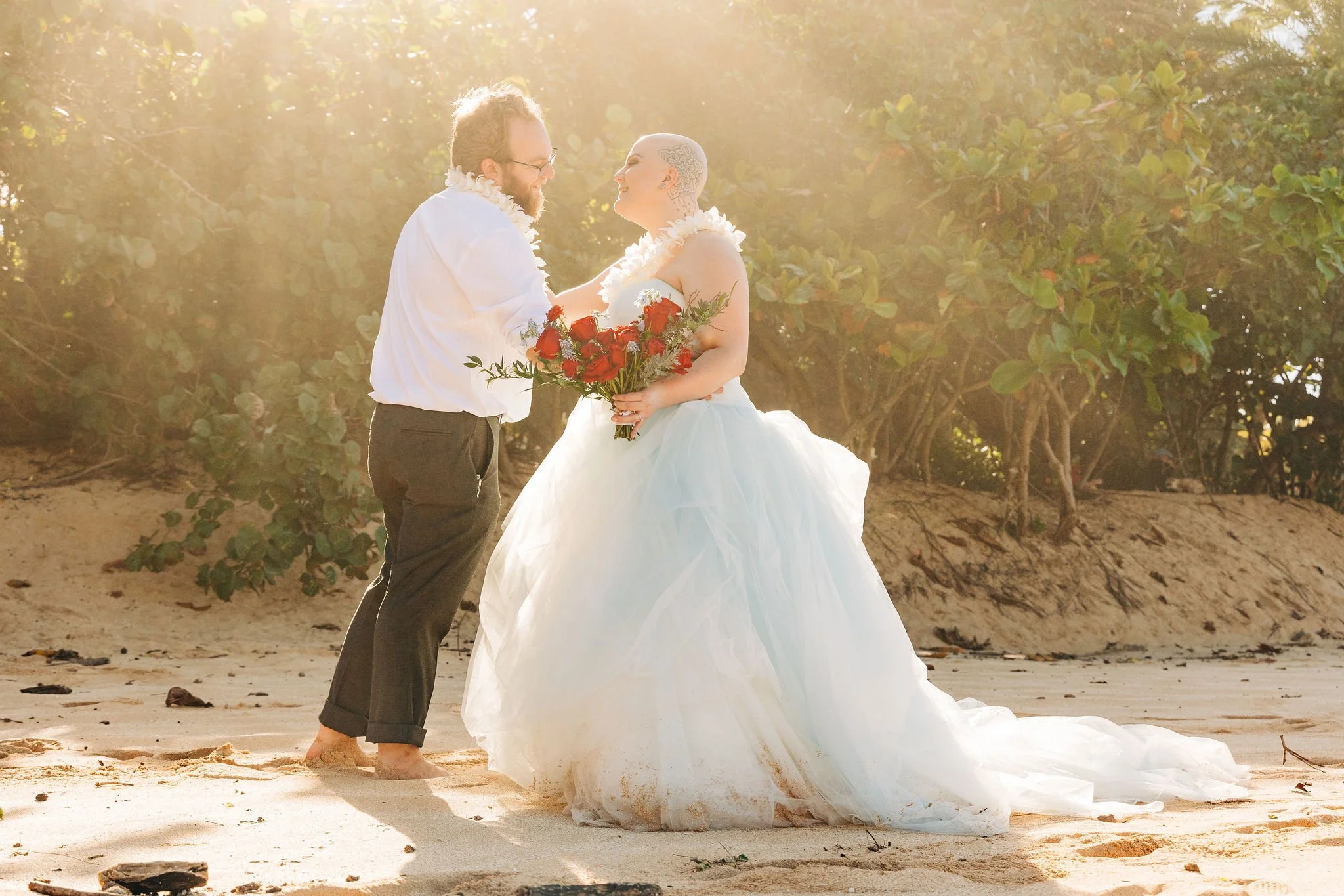 A couple in wedding attire standing on a sandy beach, holding hands and smiling at each other during sunset, surrounded by greenery.