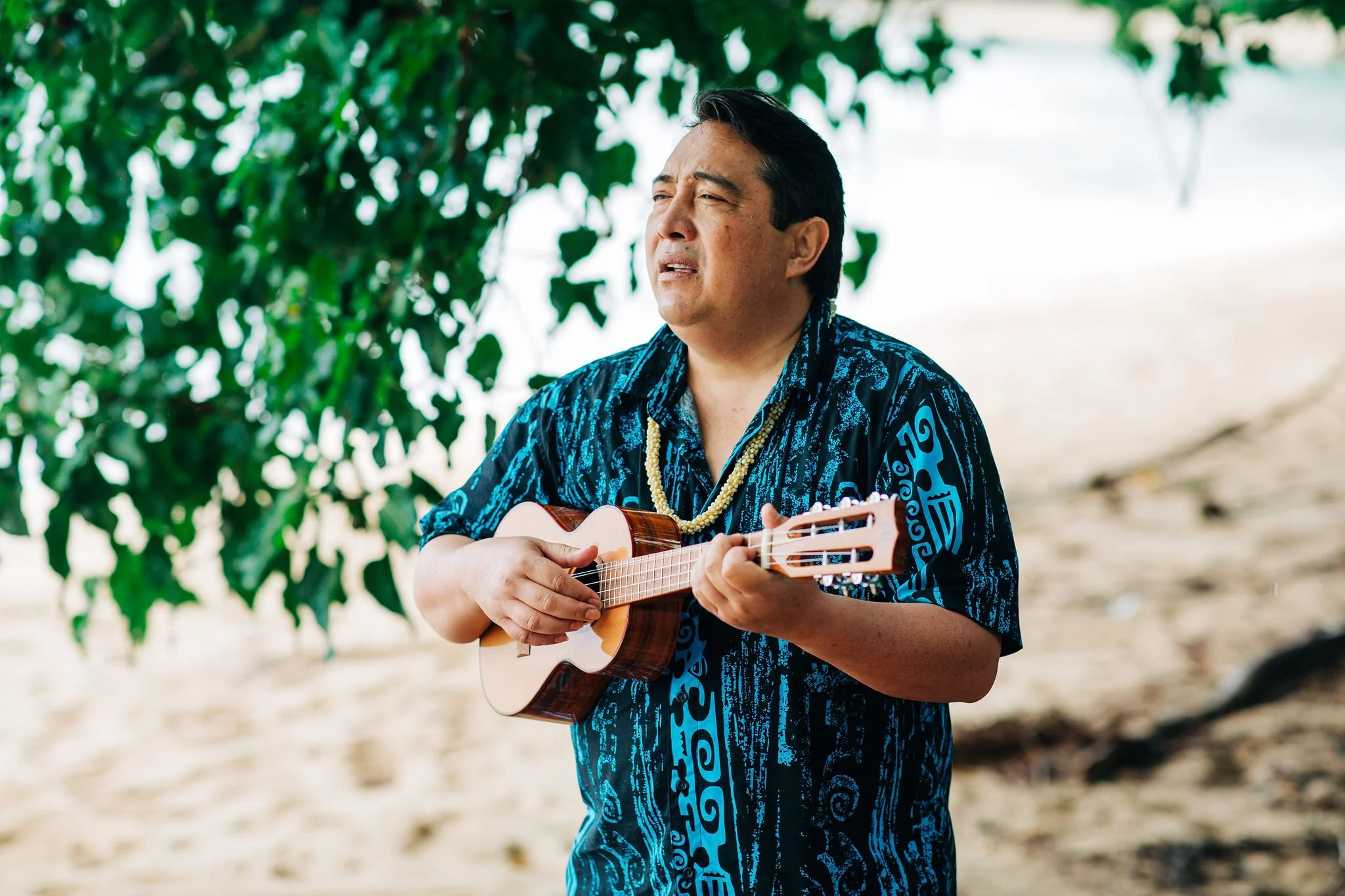 A man wearing a blue patterned shirt and a Hawai'ian lei playing a ukulele outdoors on a beach with trees in the background.