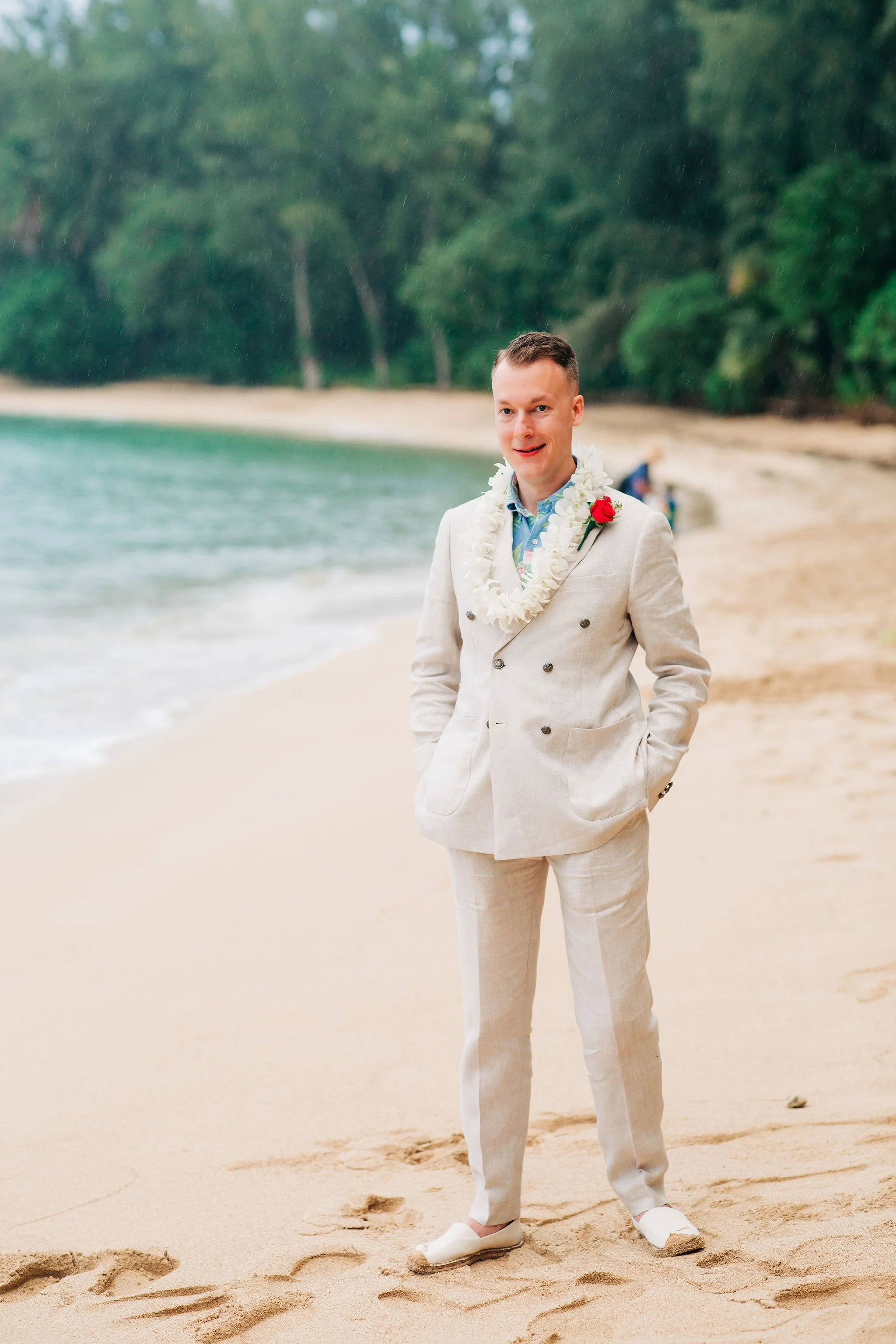 A man in a light-colored suit standing on a beach with greenery in the background. He is wearing a flower lei and a red flower on his lapel, indicating a wedding or celebration.