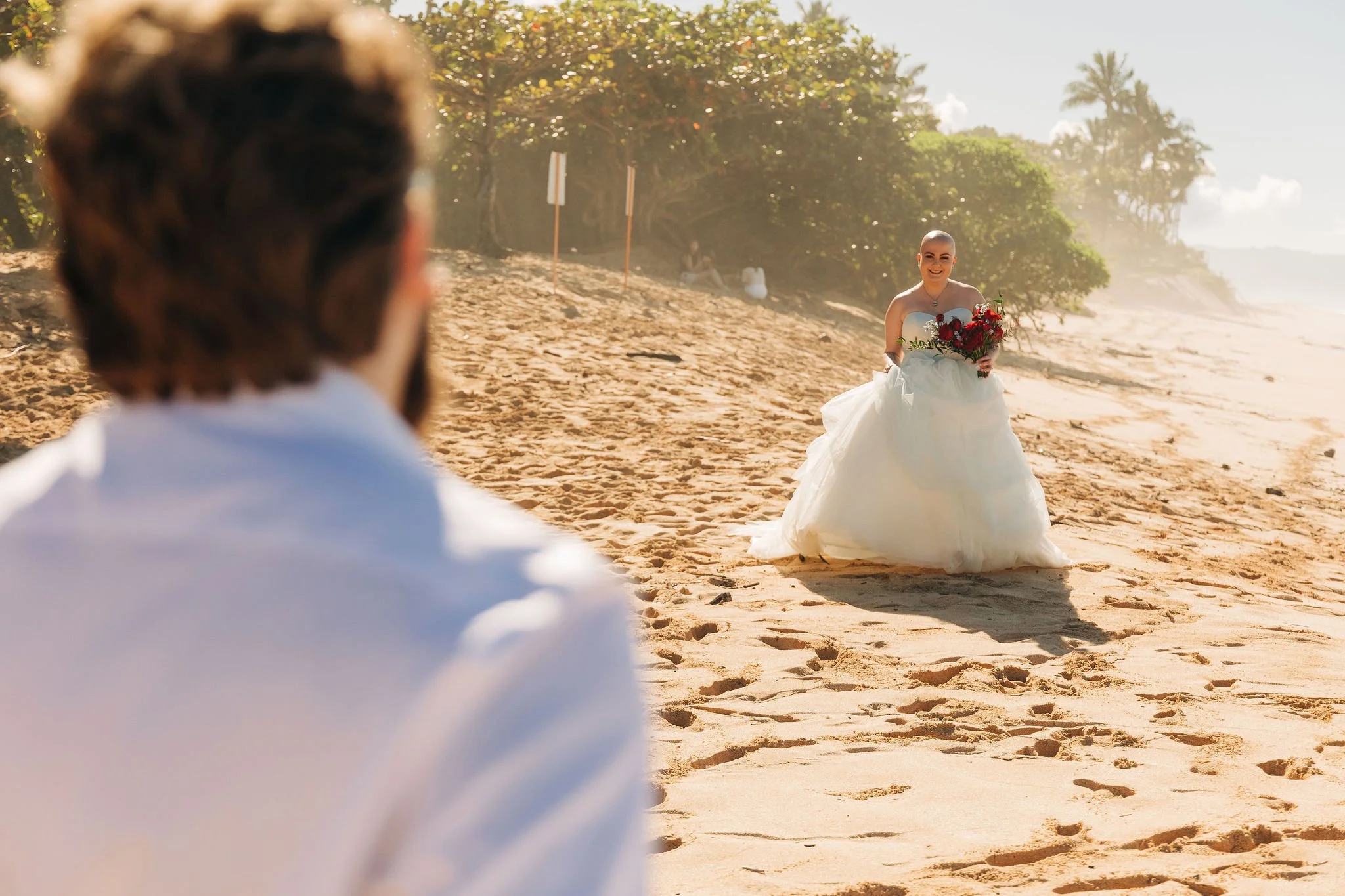 A bride in a white wedding dress holding a bouquet of red flowers walks on a sandy beach toward a man in a white shirt, with trees and a few people in the background.