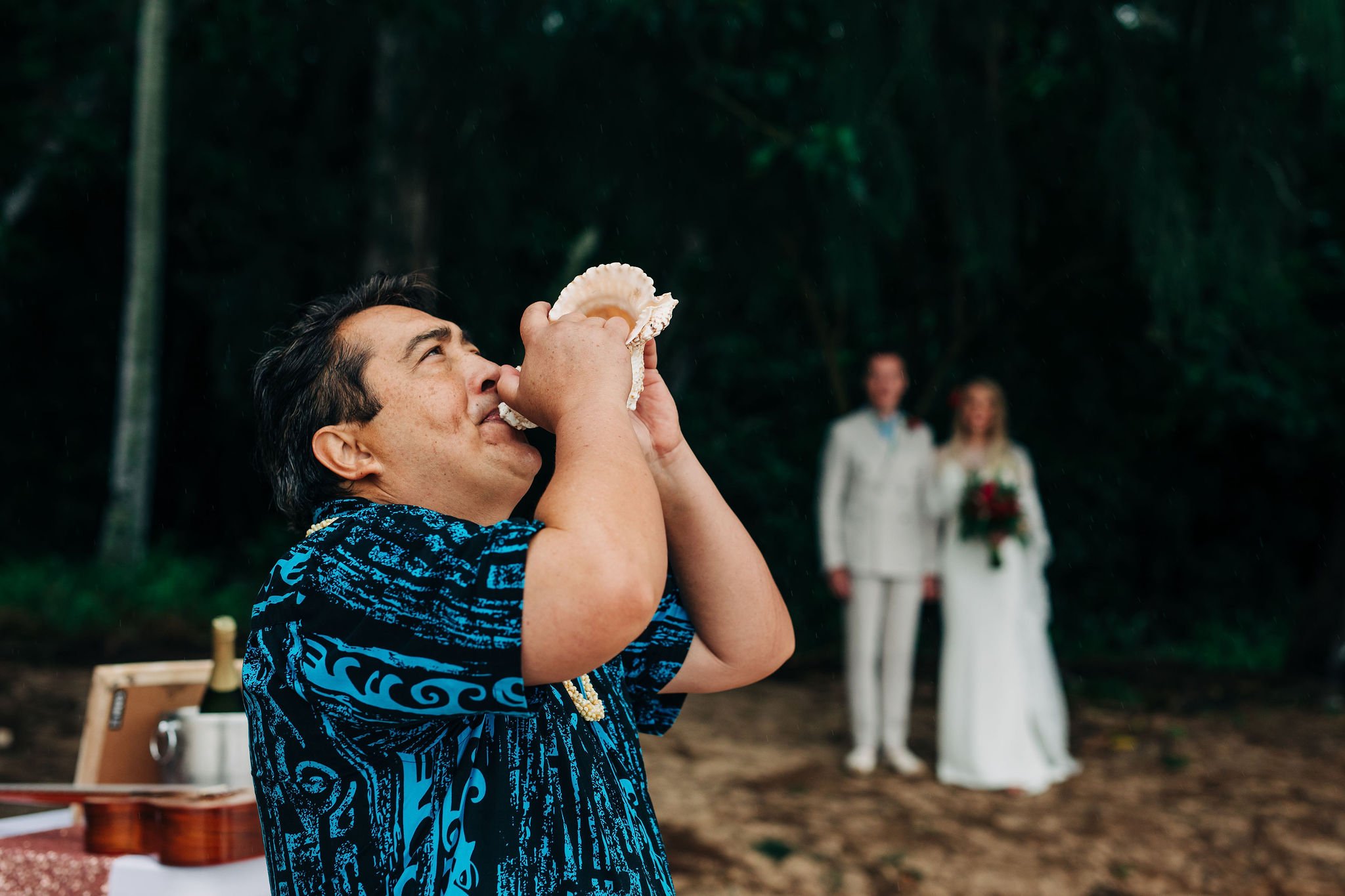 A man in a blue patterned shirt holding a seashell to his face during a celebration in an outdoor wooded area with a couple dressed in white wedding attire in the background.