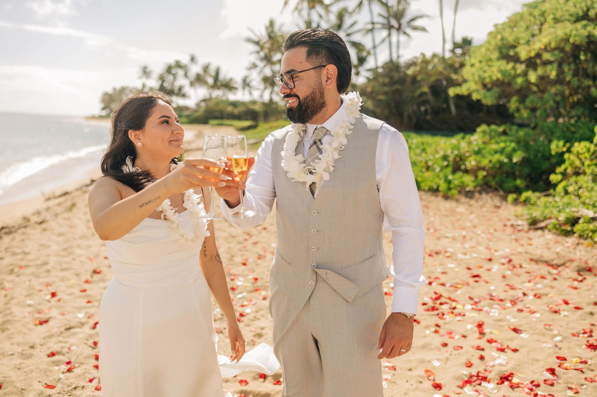 A couple in wedding attire toasting with champagne on a beach, surrounded by flower petals and lush greenery in the background.