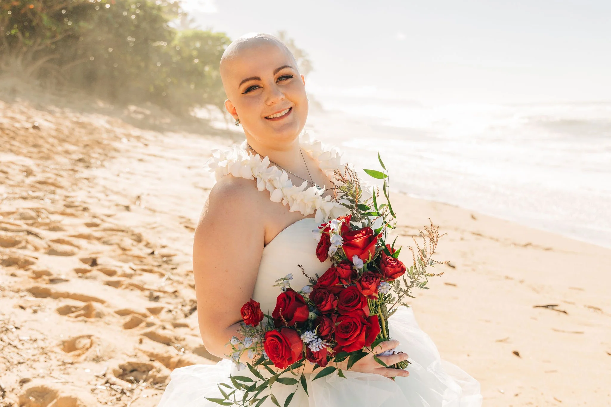 A woman wearing a white wedding dress with a floral necklace on a sandy beach, holding a bouquet of red and white flowers, smiling.