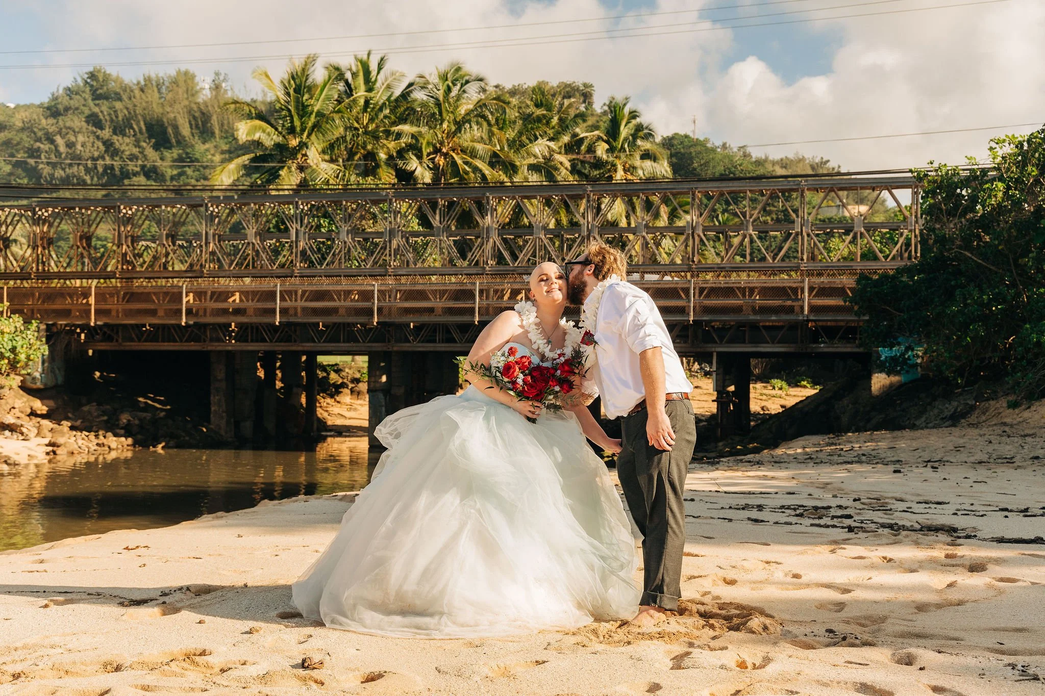 A newlywed couple on a beach, with the groom kissing the bride's temple. The bride wears a white wedding dress and holds a bouquet of red and white flowers, and the groom is dressed in a white shirt and dark pants. Behind them is a bridge, palm trees