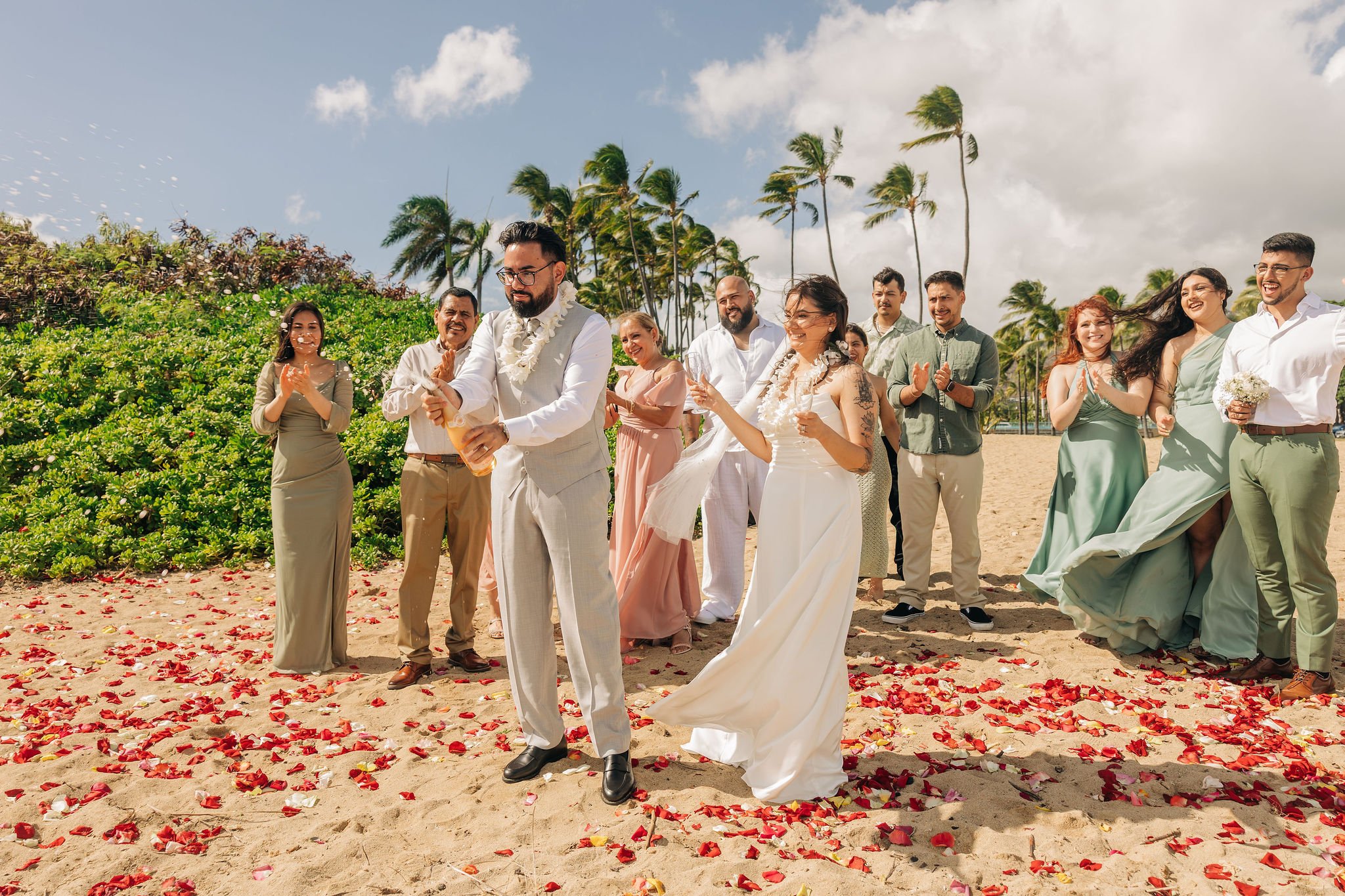 A wedding ceremony on a beach with a bride and groom surrounded by family and friends, palm trees in the background, and flower petals on the sand.