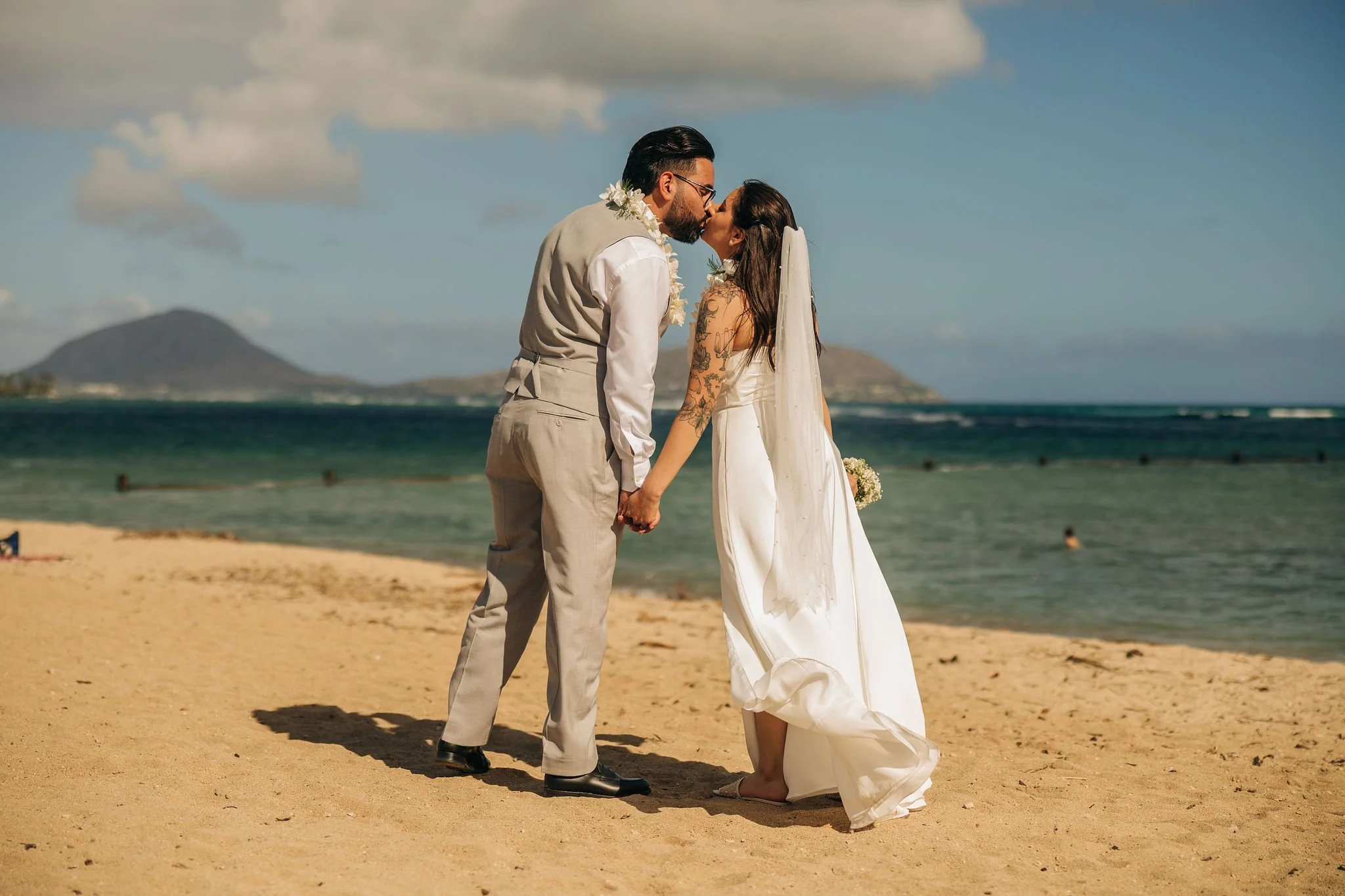 A couple in wedding attire holding hands and kissing on a sandy beach with ocean, mountains, and partly cloudy sky in the background.