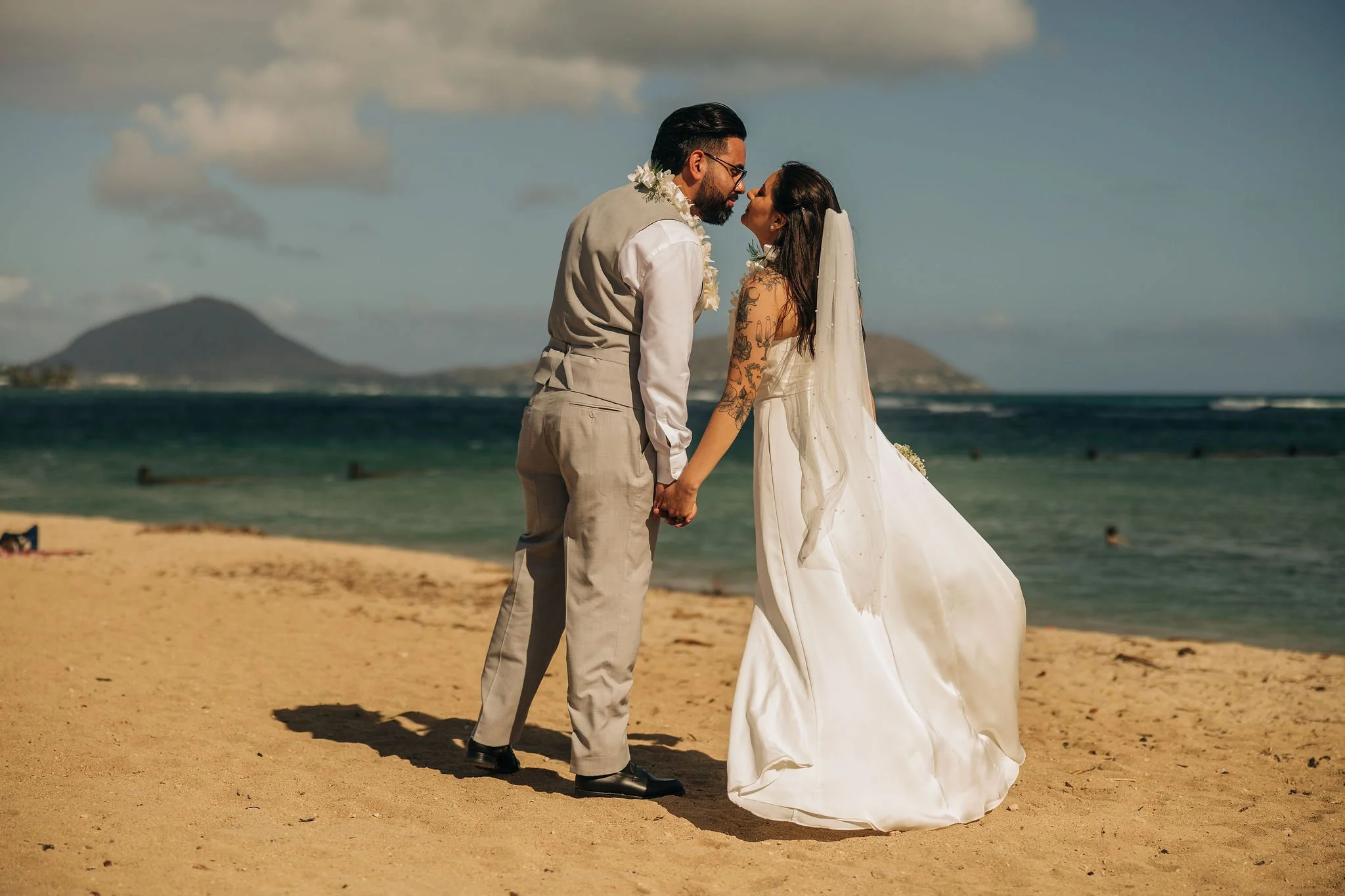 A couple in wedding attire on a sandy beach, holding hands and leaning in for a kiss, with ocean and mountains in the background.