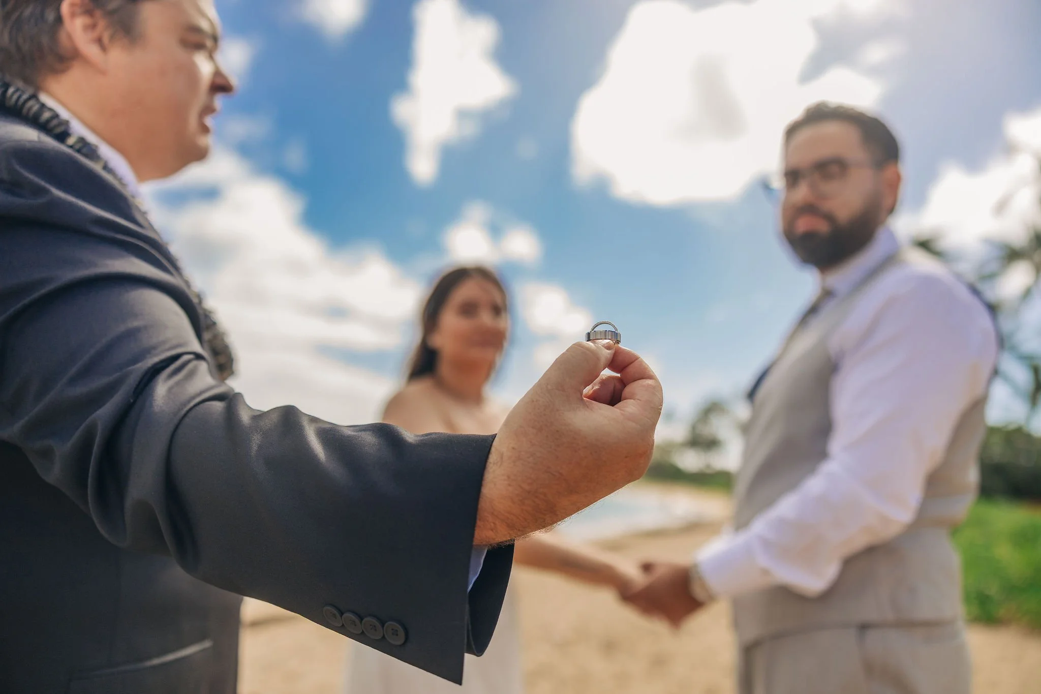 A couple holding hands outdoors on a beach, during their wedding ceremony. The officiant is holding a wedding ring in the foreground.