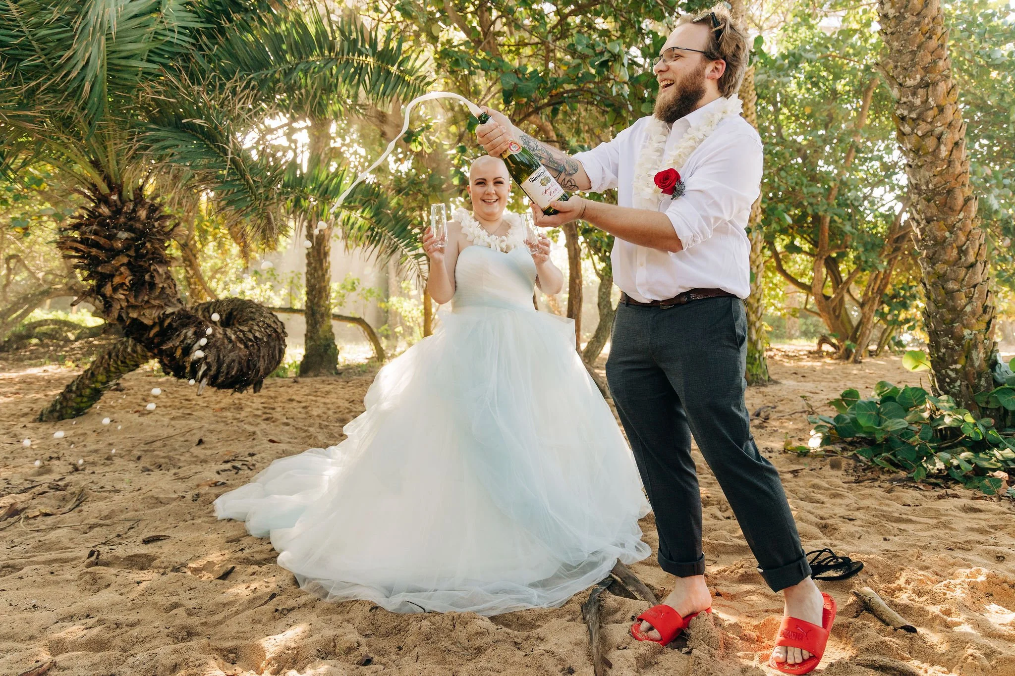 A newlywed couple celebrating on the beach, with the groom popping open a champagne bottle and the bride holding two glasses, both smiling and wearing wedding attire surrounded by palm trees.