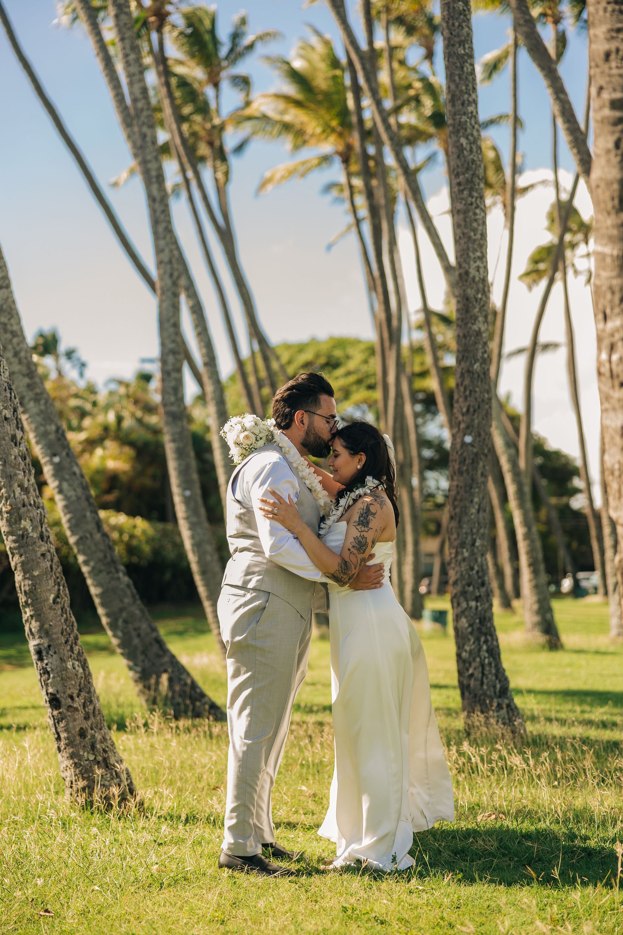 A couple dressed in wedding attire embracing each other in a tropical outdoor setting with palm trees and blue sky.
