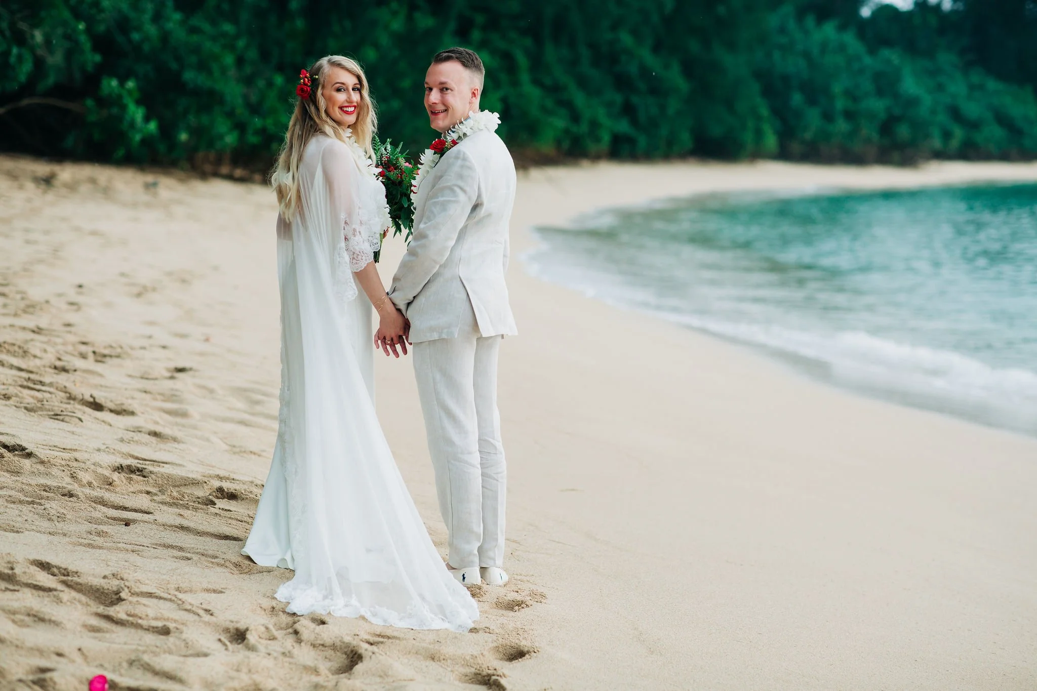 A bride and groom holding hands on a sandy beach with green trees in the background, dressed in wedding attire with the bride smiling, holding a bouquet of flowers.