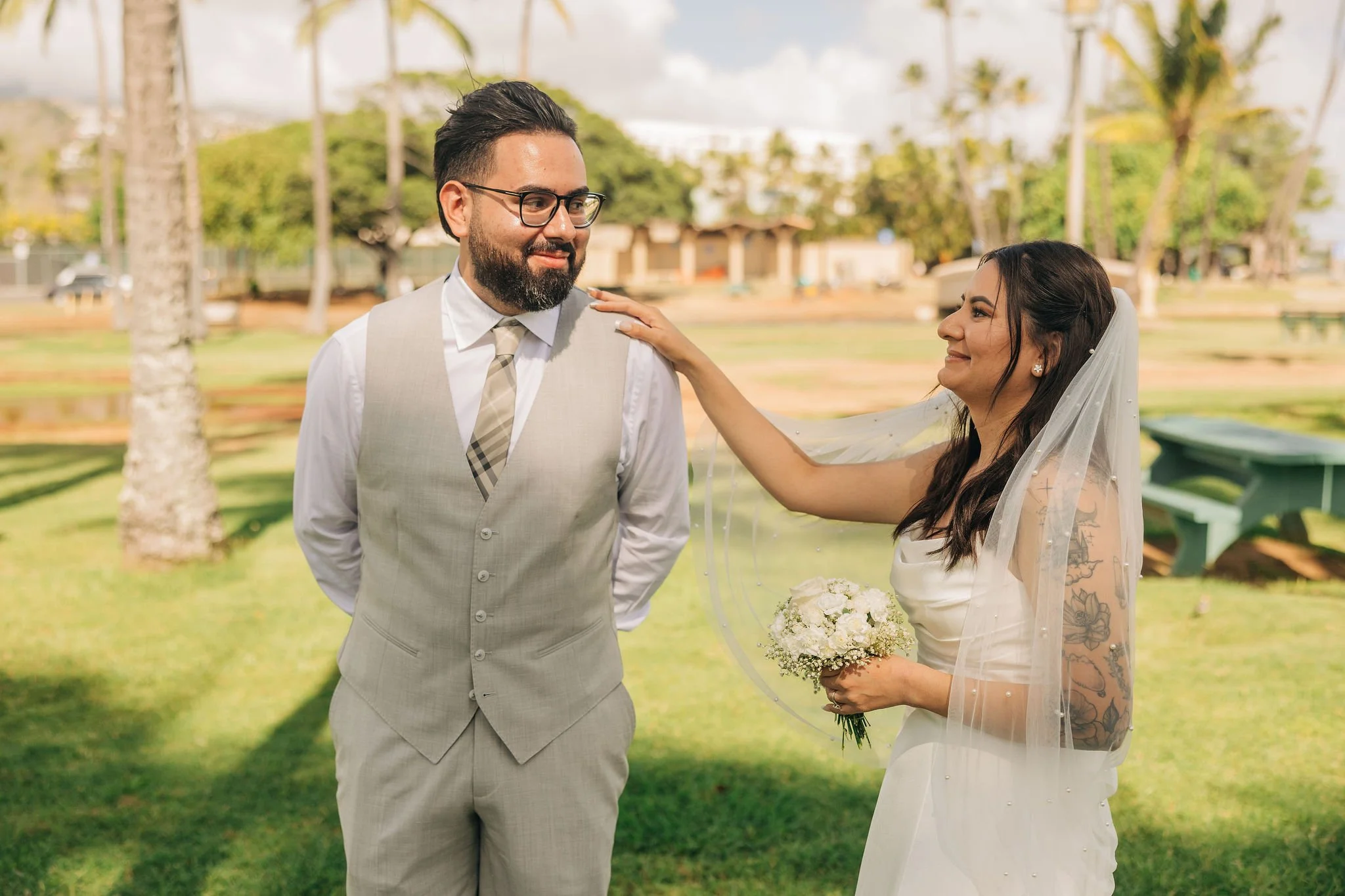 A groom and bride standing outdoors, smiling at each other. The groom has a beard, glasses, and is dressed in a light gray vest with a white shirt and tie. The bride has long dark hair with a veil, and tattoos on her arm, holding a bouquet of white f