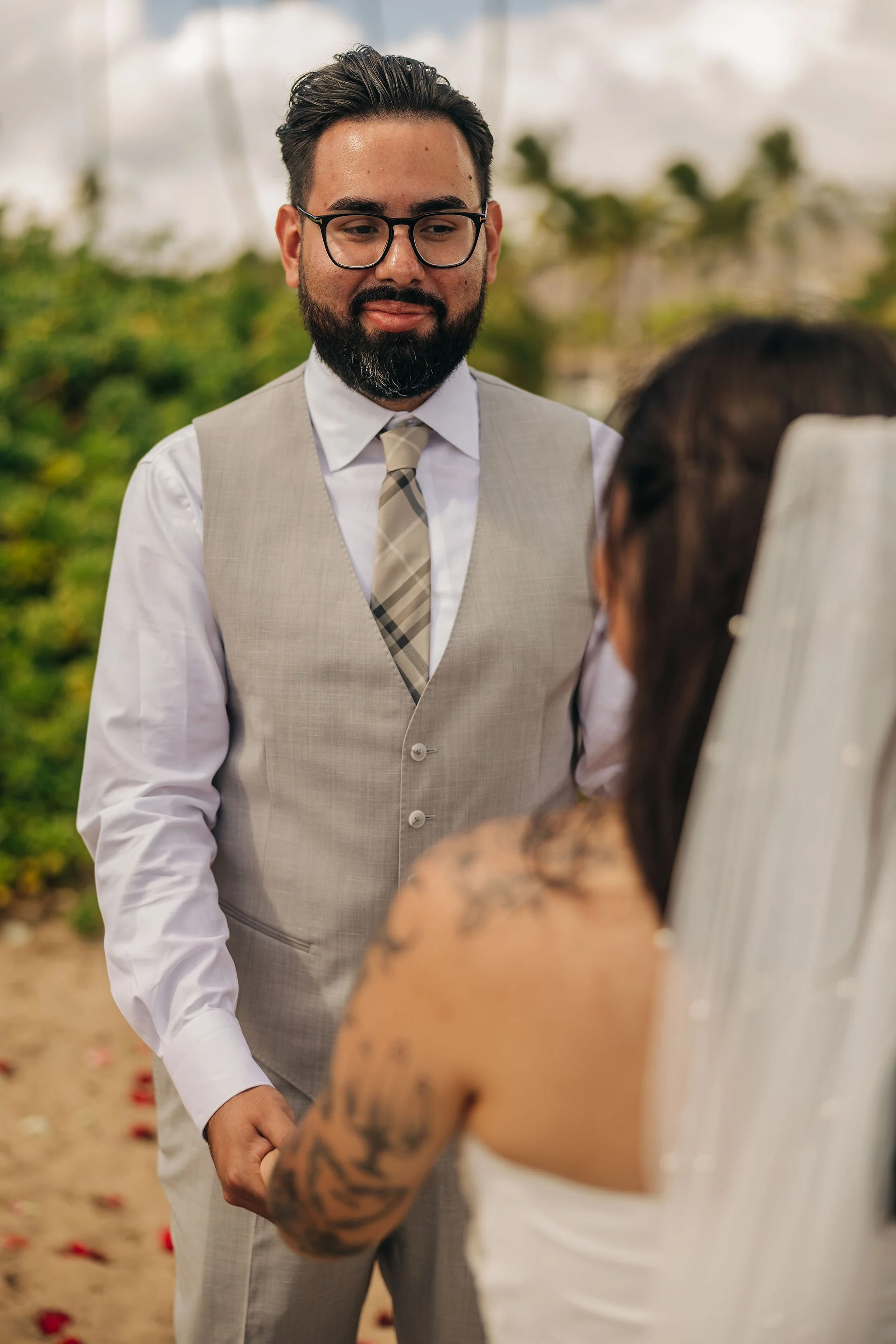 Groom in a light gray vest, white shirt, and plaid tie standing outdoors, looking at bride with long dark hair and tattoos, on a beach with green foliage in background.
