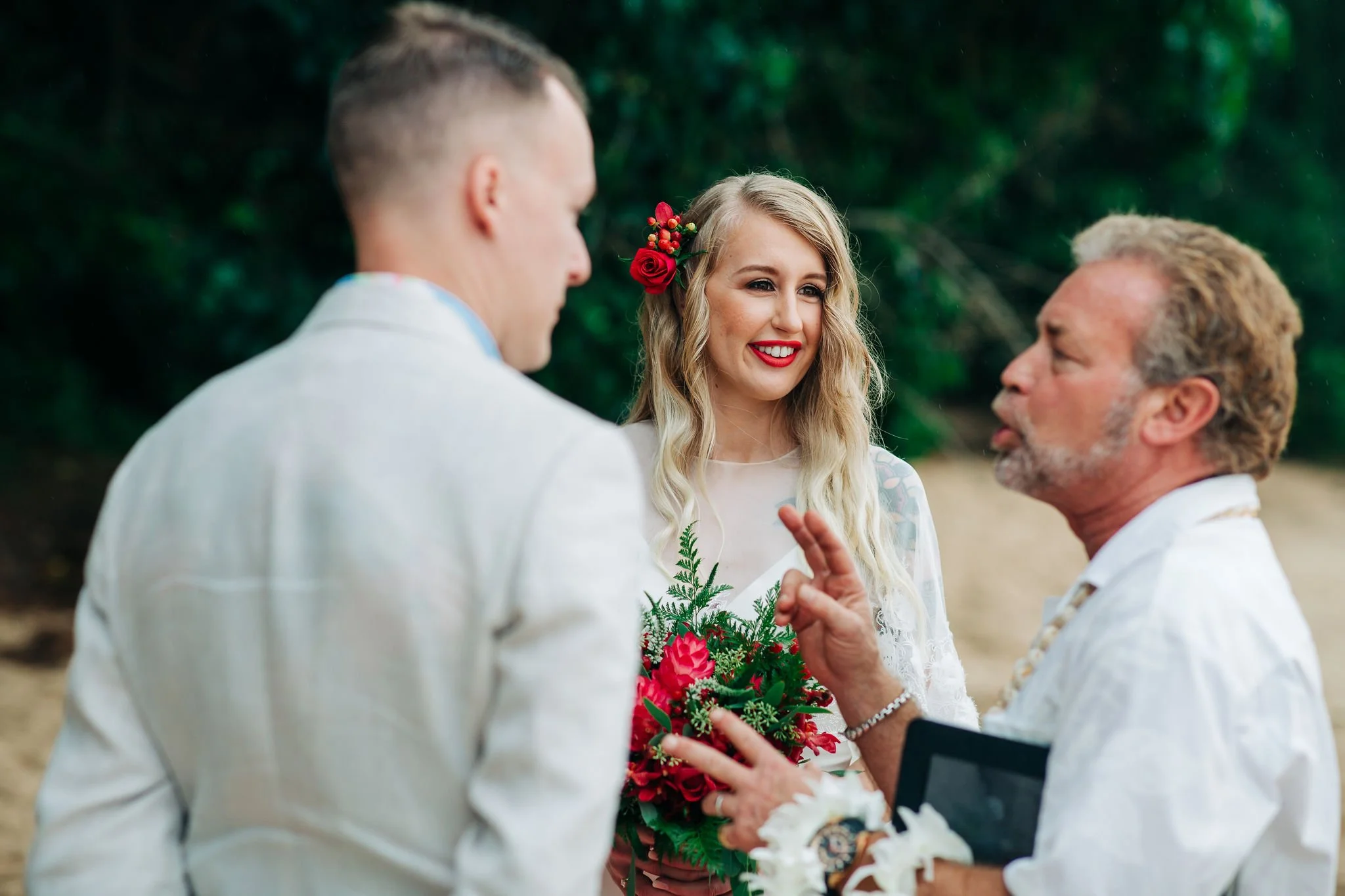 A woman with long blond hair and a red flower in her hair, smiling with red lipstick, holding a bouquet of red flowers and greenery, standing between two men in white suits in an outdoor setting with green foliage in the background.