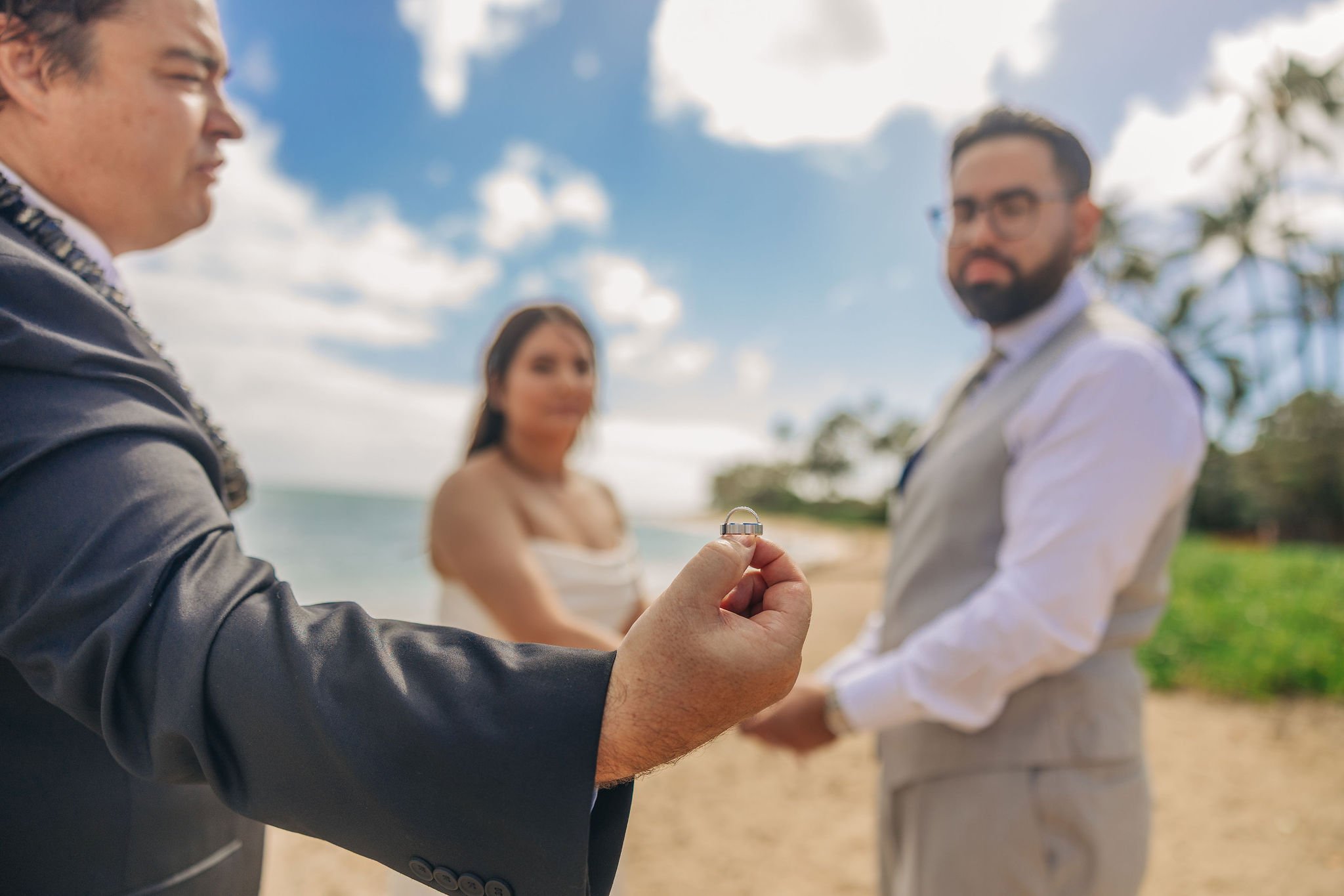 A man in a black suit holds a wedding ring in the foreground with two people, a woman and a man in formal attire, standing behind him on a beach.
