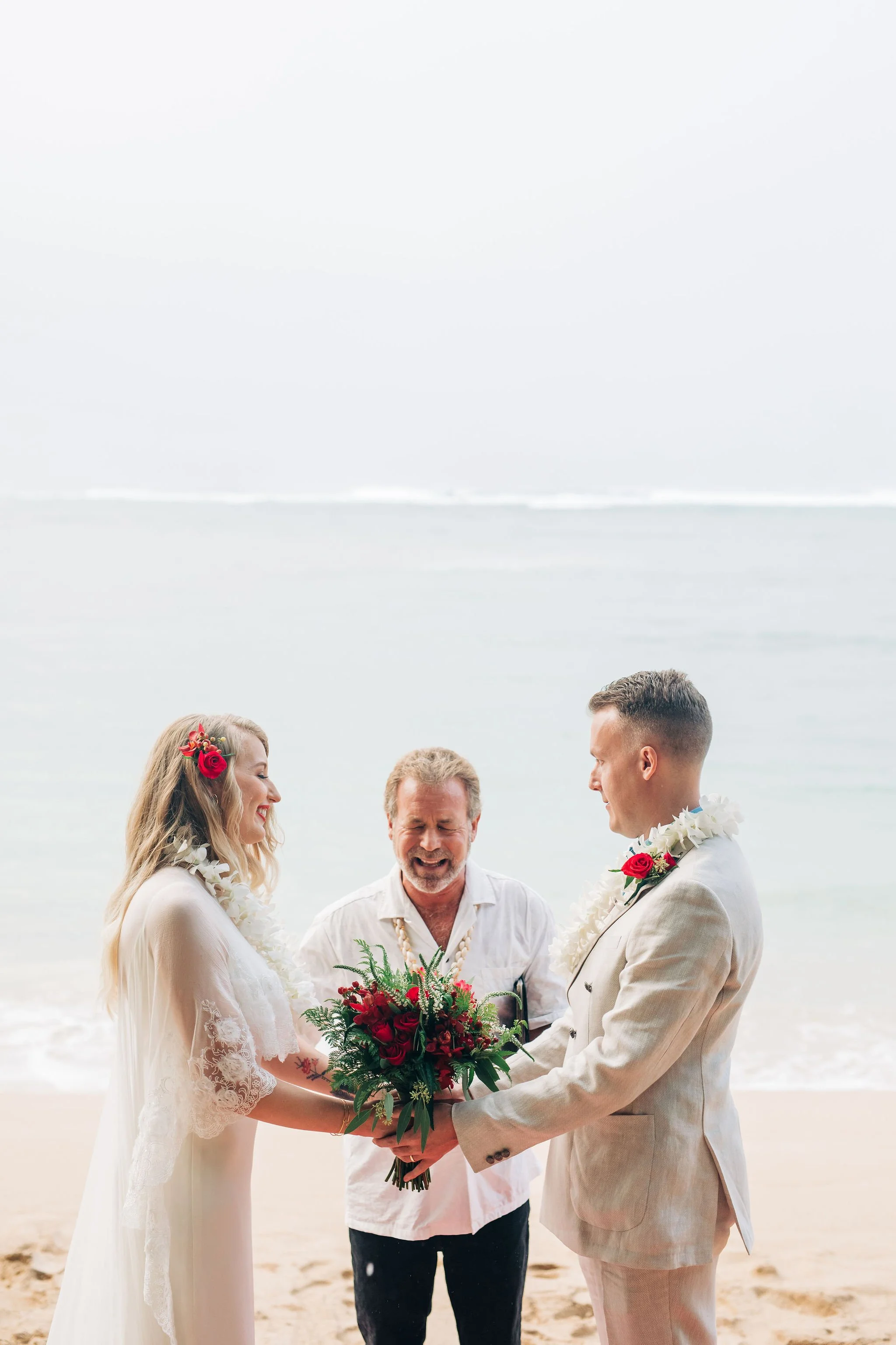 A couple getting married on the beach with an officiant, all holding a bouquet of red flowers. The bride has blonde hair with floral accessories, and the groom has short hair. The officiant is smiling, and the sea is in the background.