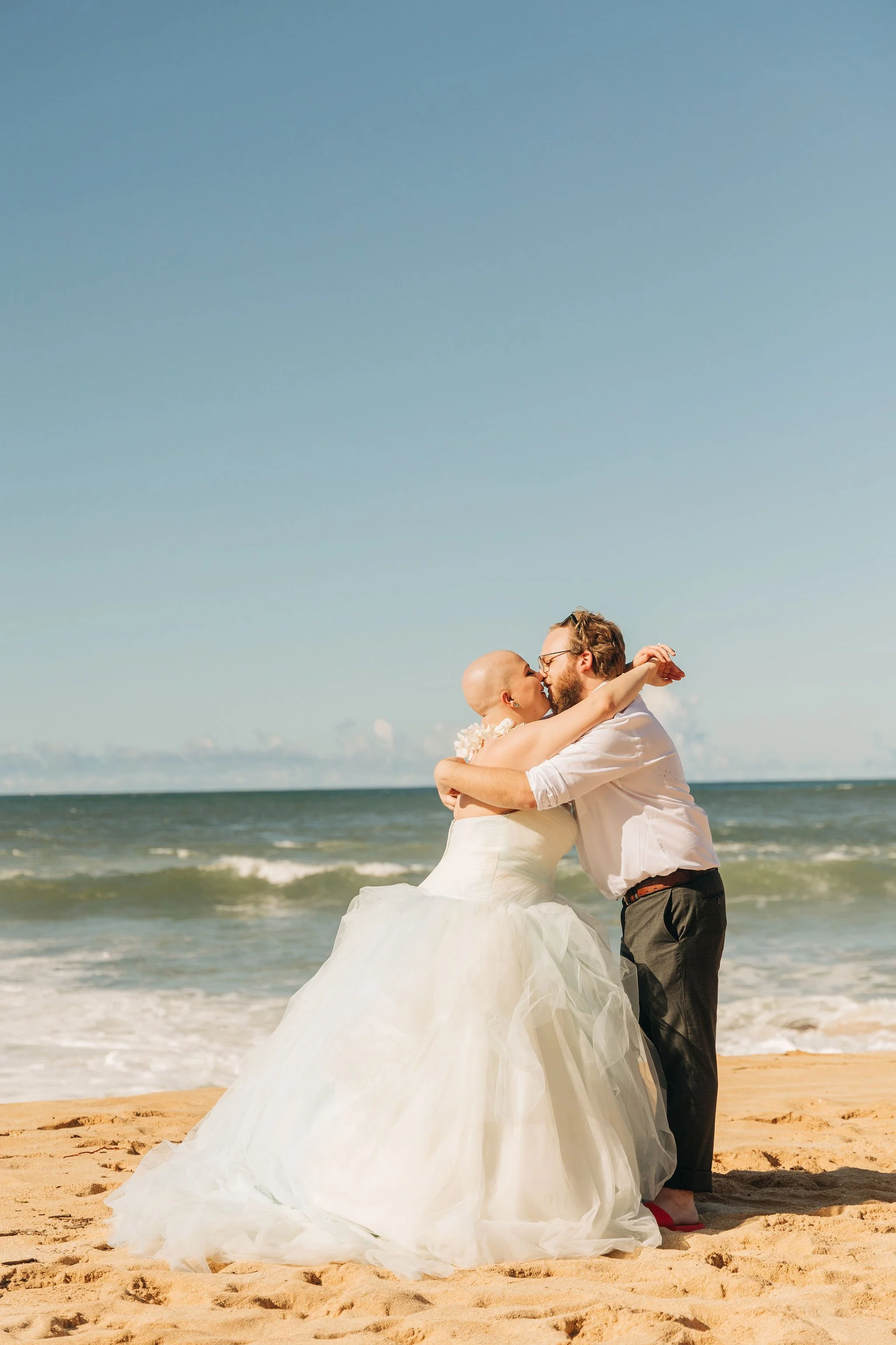 A couple dressed in wedding attire embracing and kissing on a sandy beach with the ocean and blue sky in the background.