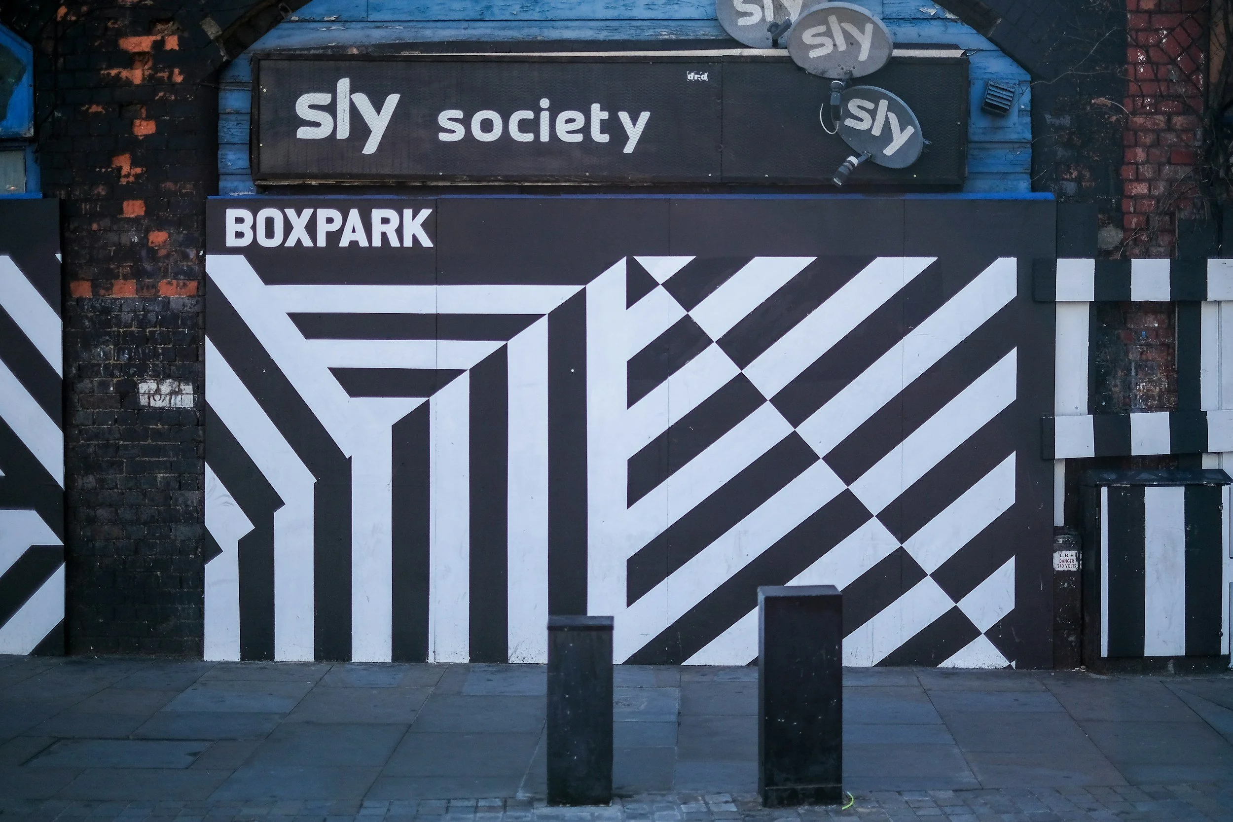 Street art mural with black and white geometric stripes and the word 'BOXPARK' on the left. Above it, a black sign reads 'sky society' with three satellite dishes labeled 'sky' mounted above.