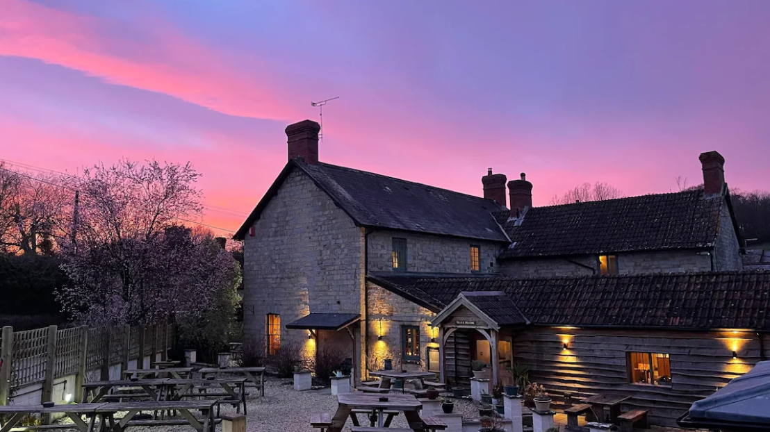 A rustic stone building and a wooden structure outside with outdoor tables, set against a colorful sunset sky with purple and pink hues, and blooming trees.