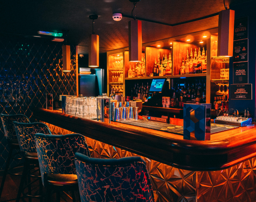 A dimly lit bar with a polished wooden counter, glassware, and bottles of liquor displayed on illuminated shelves behind the bar. Bar stools with patterned upholstery are lined along the counter, and there are various signs and equipment on the bar.