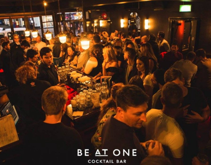 Crowded cocktail bar with many patrons standing and socializing, warm lighting, and bartenders serving drinks.