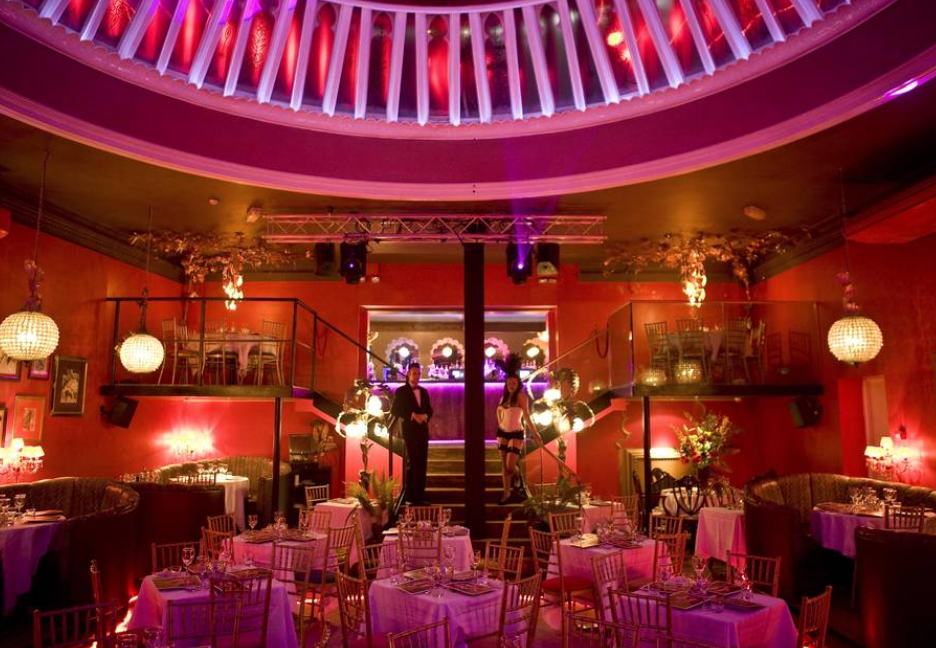 Upscale restaurant interior with pink lighting, round tables set for dining, and a second-floor balcony with additional seating overlooking the main area.