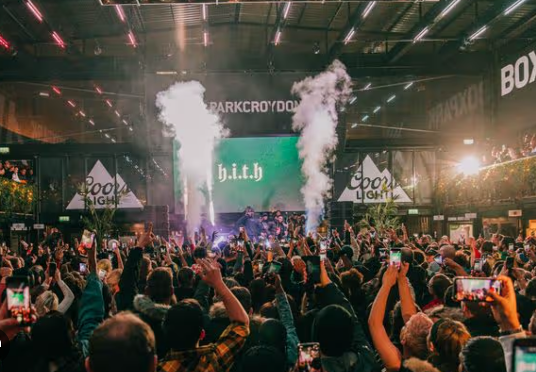 Crowd at an indoor concert or live event, with many people raising their phones to record or take photos of the stage. The stage has performers, smoke effects, and a large screen displaying the text 'H.i.t.h'. Bright stage lights and advertising banners, including 'Coors Light,' are visible.