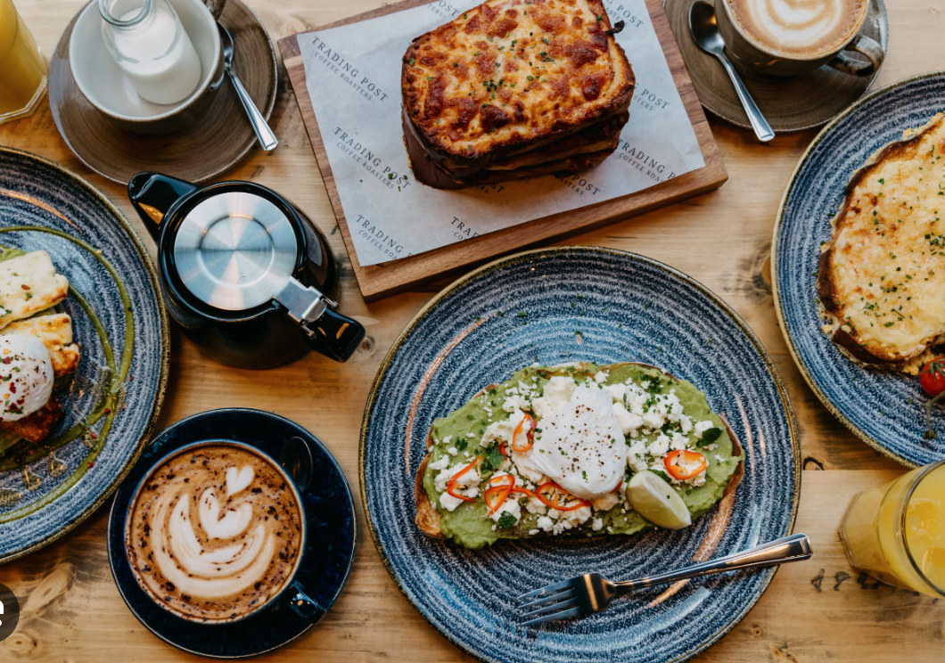 A top-down view of a breakfast or brunch spread on a wooden table with various dishes including toast with avocado, poached egg, and toppings, a slice of lasagna or baked casserole, a cup of coffee with foam, a cup of cappuccino with latte art, a small glass of orange juice, a teapot, and cream or milk in a small pitcher.