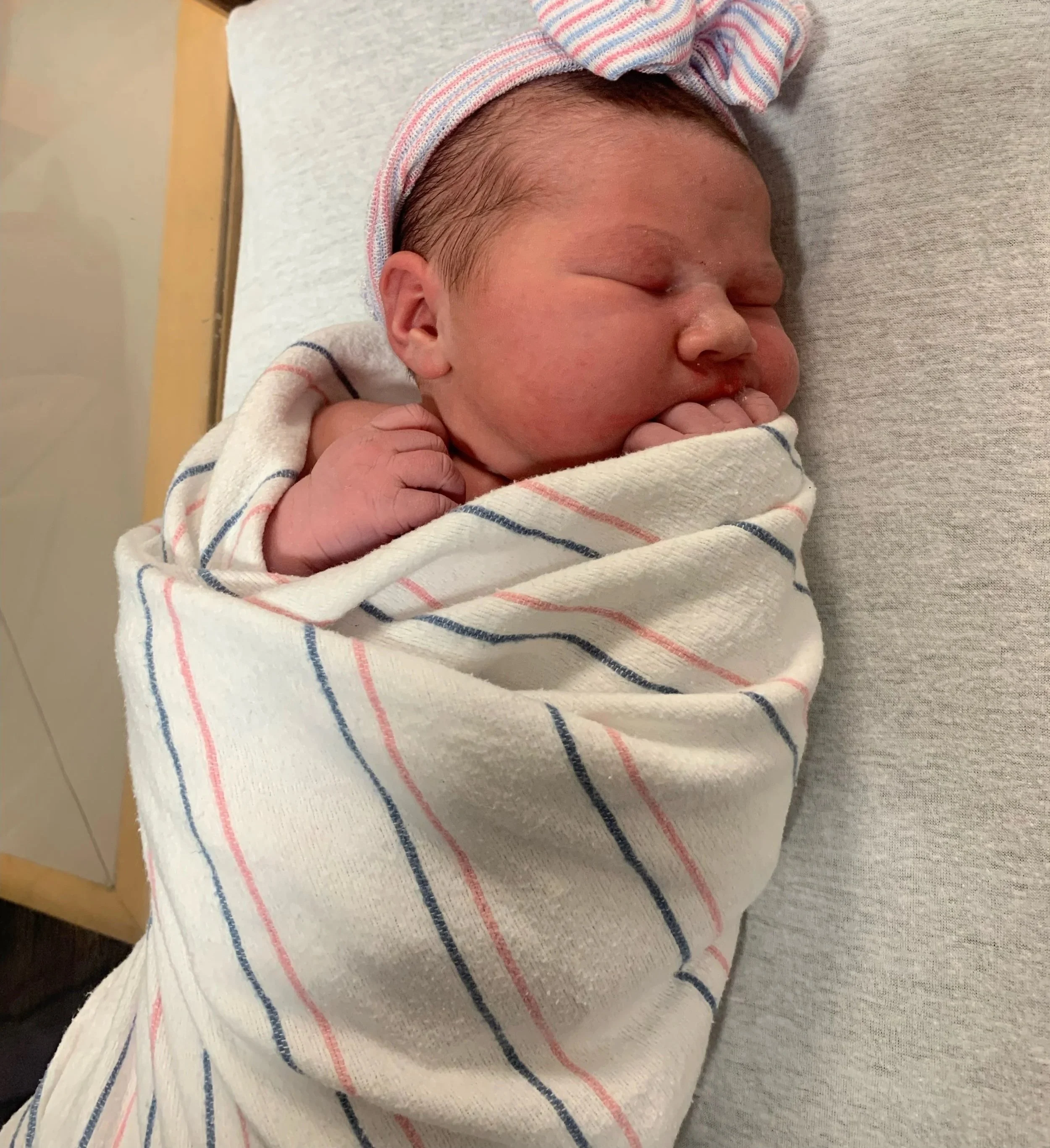 Newborn baby wrapped in a hospital blanket with pink and blue stripes, wearing a matching striped hat, sleeping peacefully on a hospital bed.