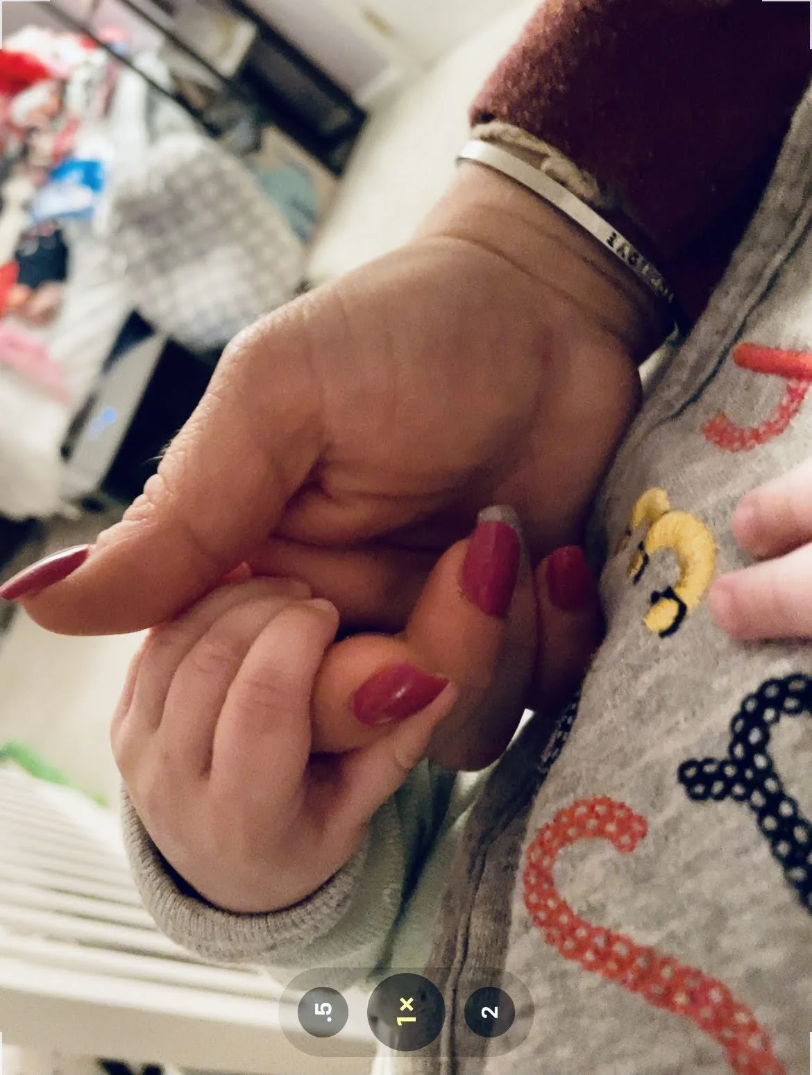Close-up of two people holding hands, one with red painted nails and the other with unpainted nails, against a background of a cluttered room.