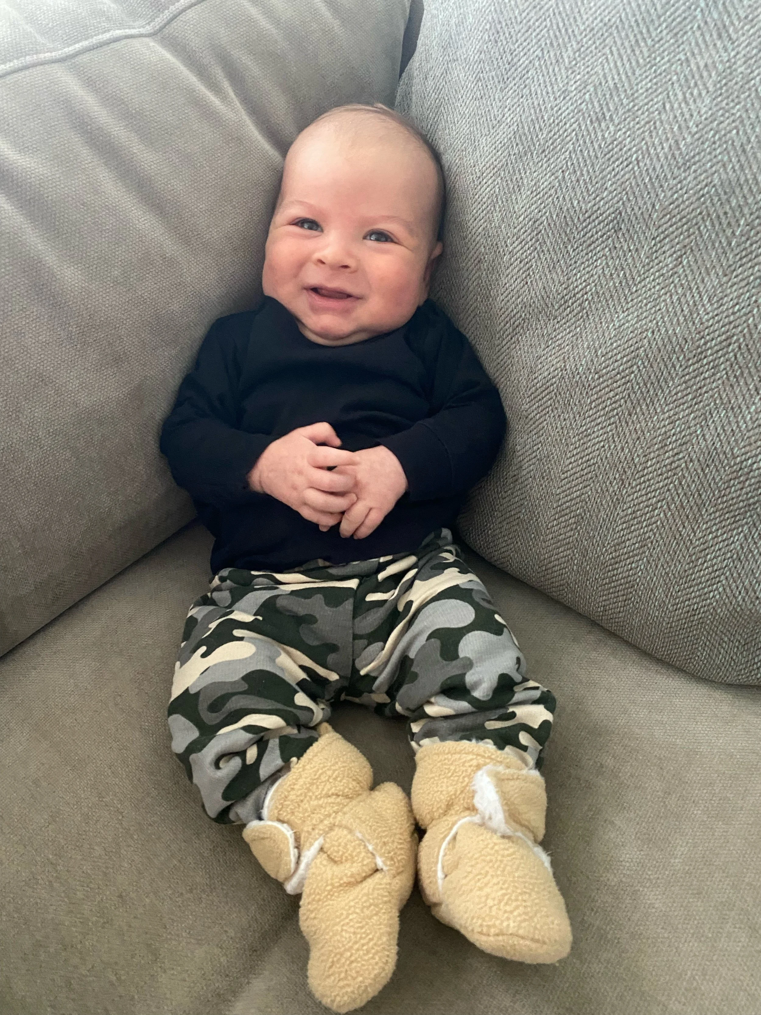 A smiling baby lying on a couch, wearing yellow boots, camo pants, and a black shirt.