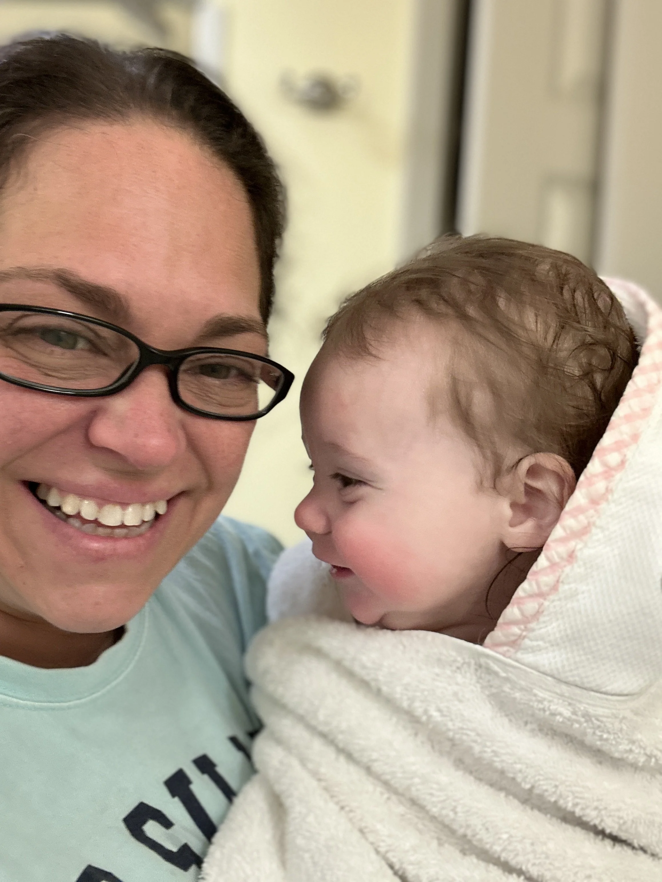 A woman with glasses smiling and holding a baby wrapped in a white towel, both looking at each other with affection.