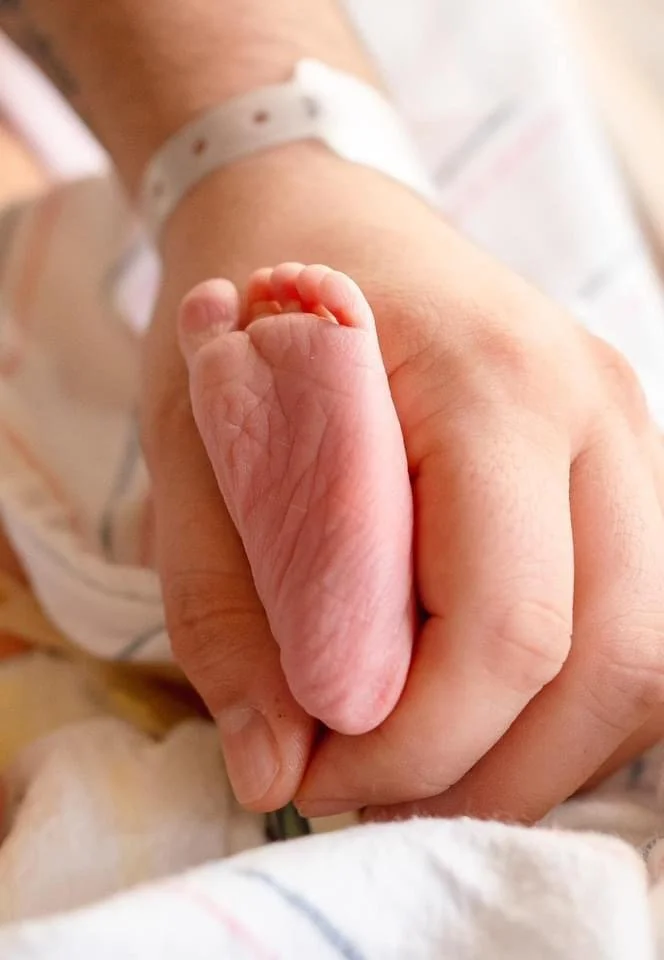 Close-up of a newborn baby's tiny hand gripping an adult's finger, with hospital wristbands visible.